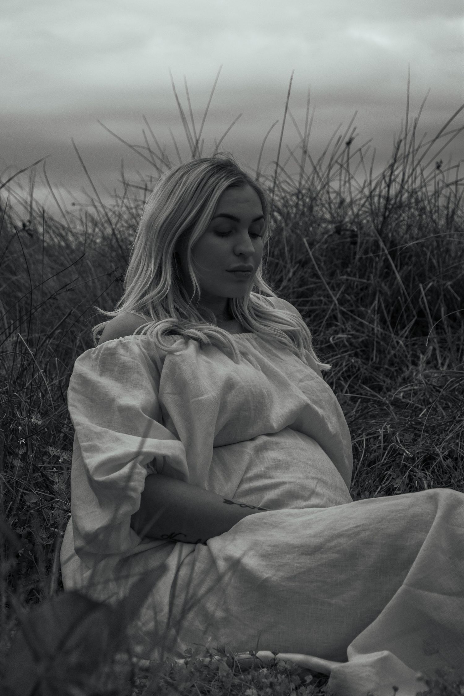 A dreamy black and white maternity photo of a woman in a white dress reclining among tall grasses.