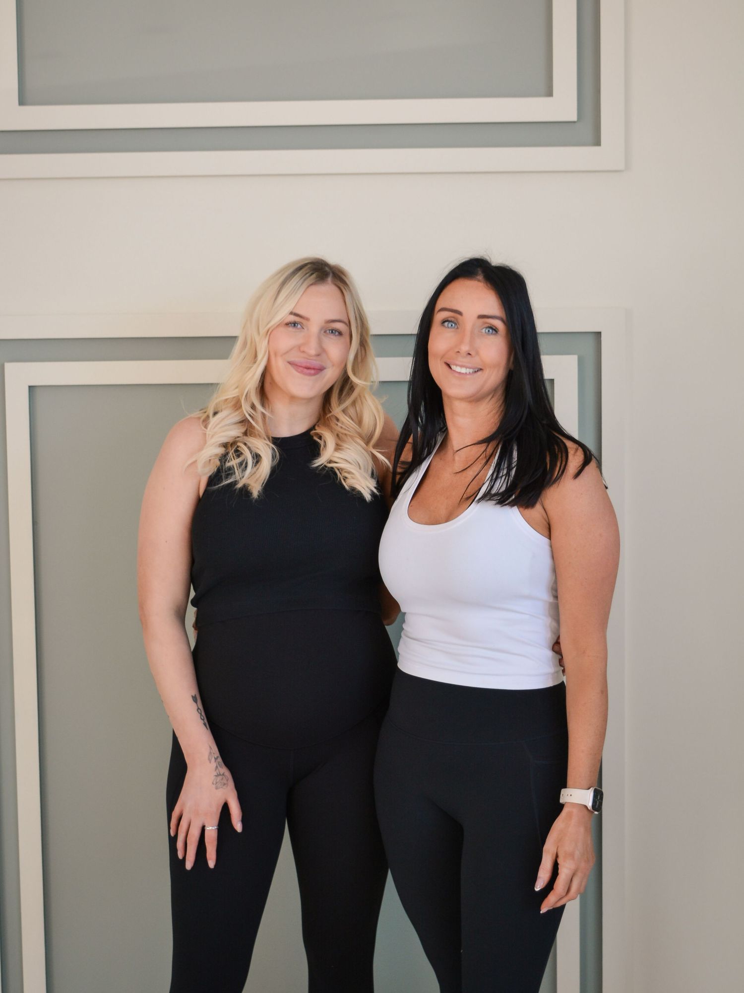 Two women in athletic wear posing together against a light colored wall.