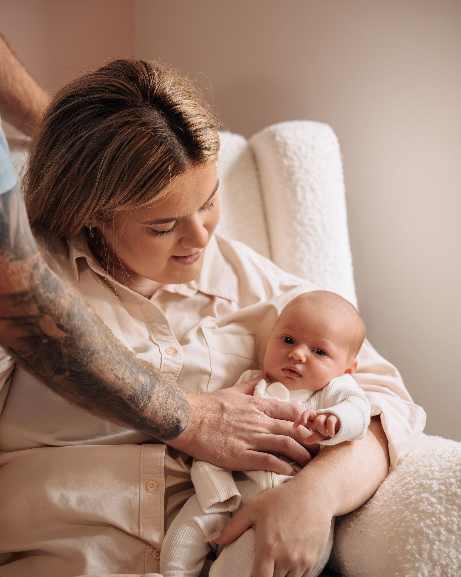 A mother tenderly holds her newborn baby while seated in a cozy white chair, with a tattooed arm visible nearby.
