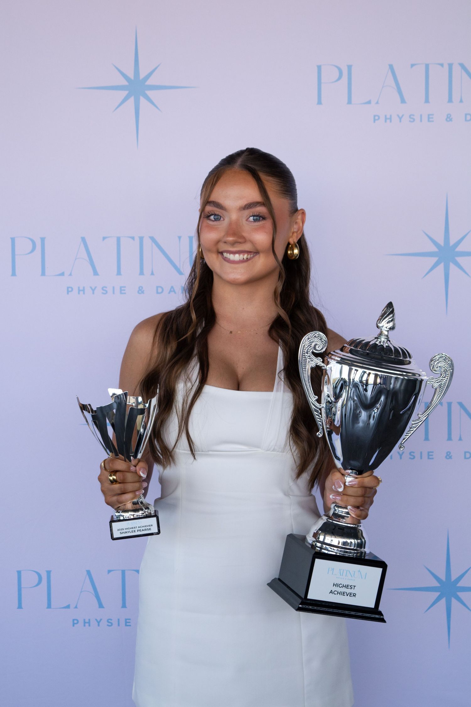 A person in white dress holds two silver trophy cups against a branded backdrop.