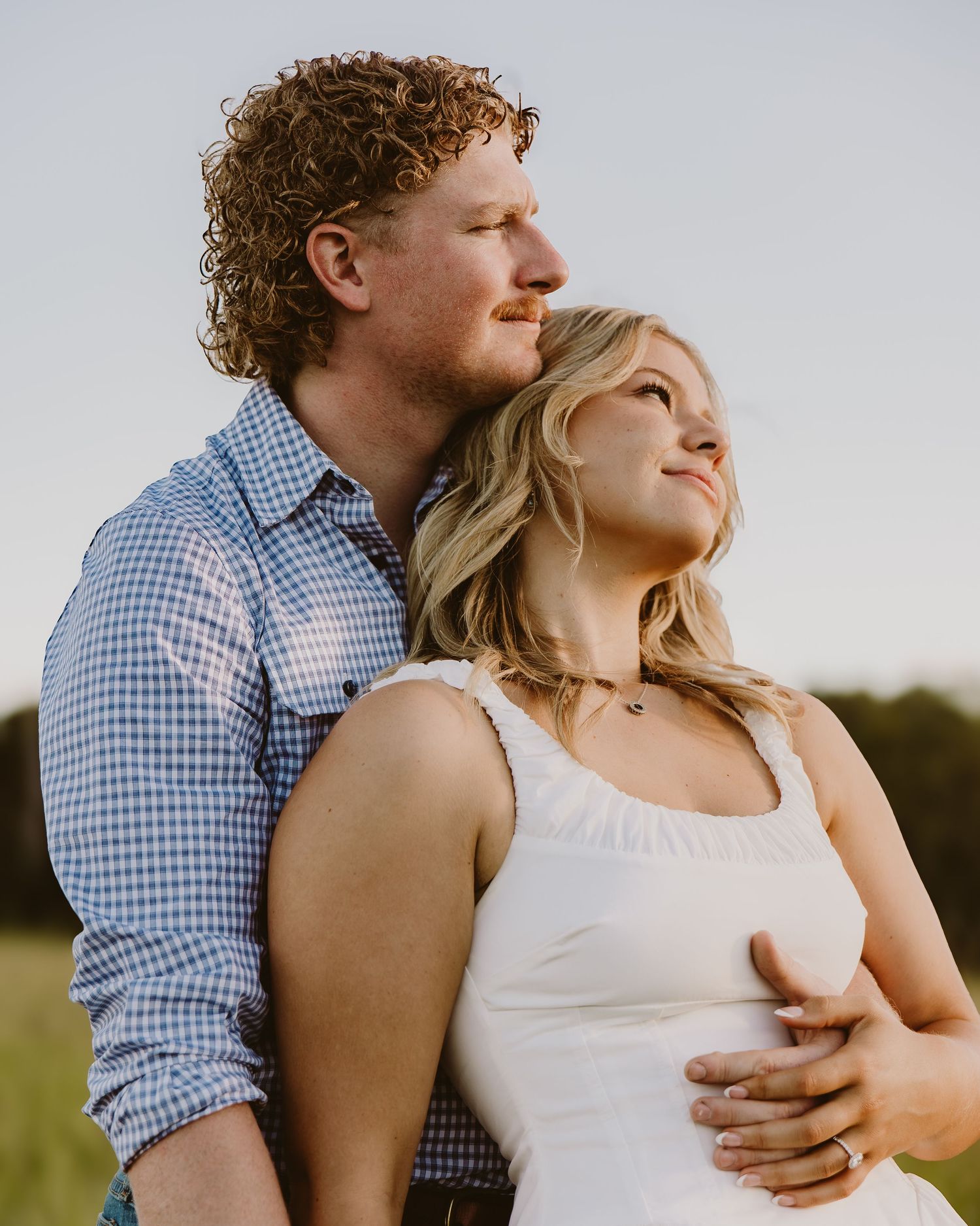 A couple embraces outdoors at golden hour, man with curly red hair holds blonde woman in white dress from behind.