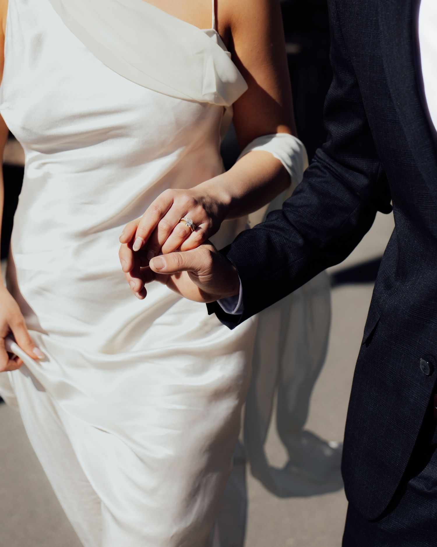 Close-up of bride and groom holding hands, showcasing wedding ring in elegant white bridal gown.