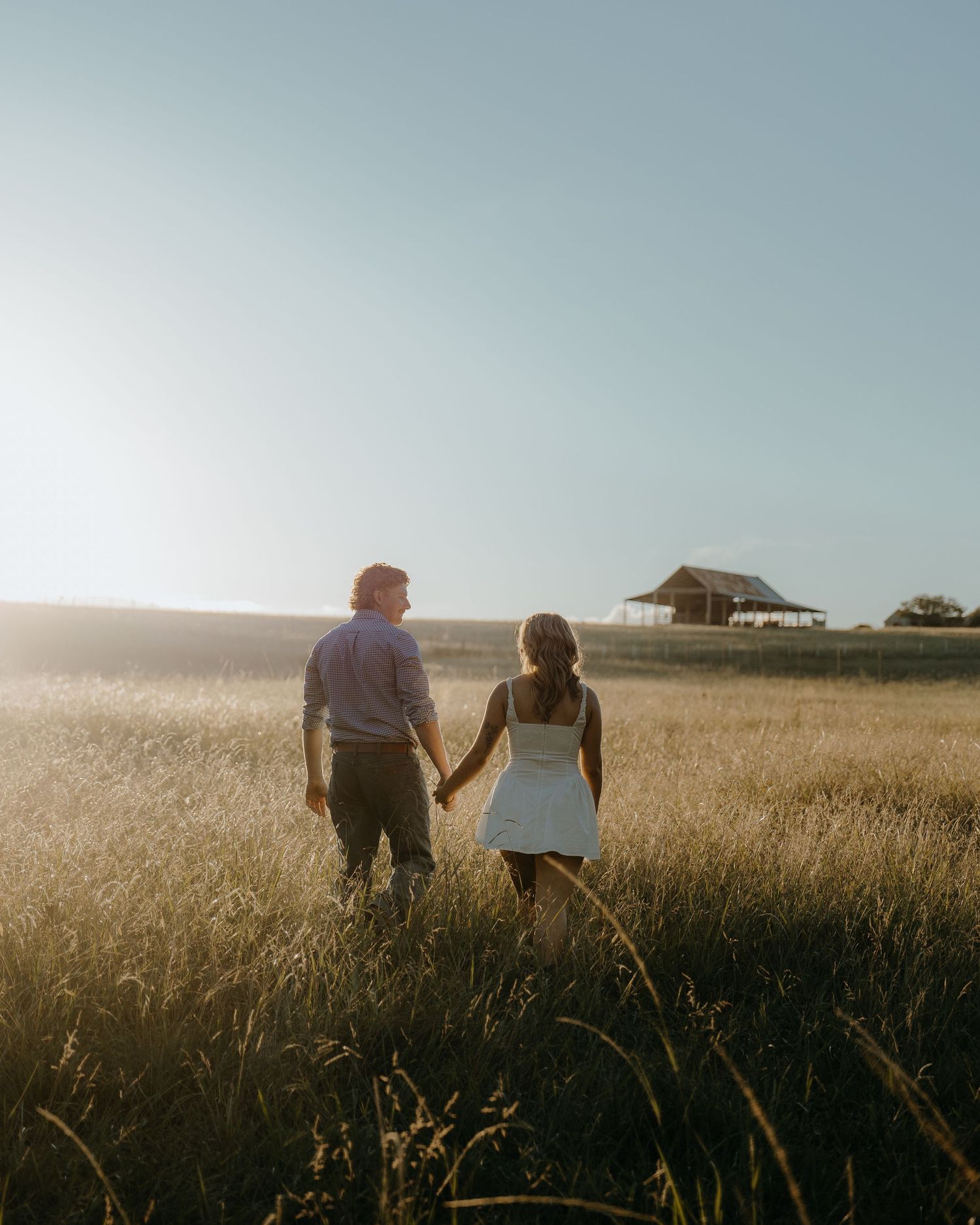 Couple holding hands walking through golden field toward rustic barn at sunset.