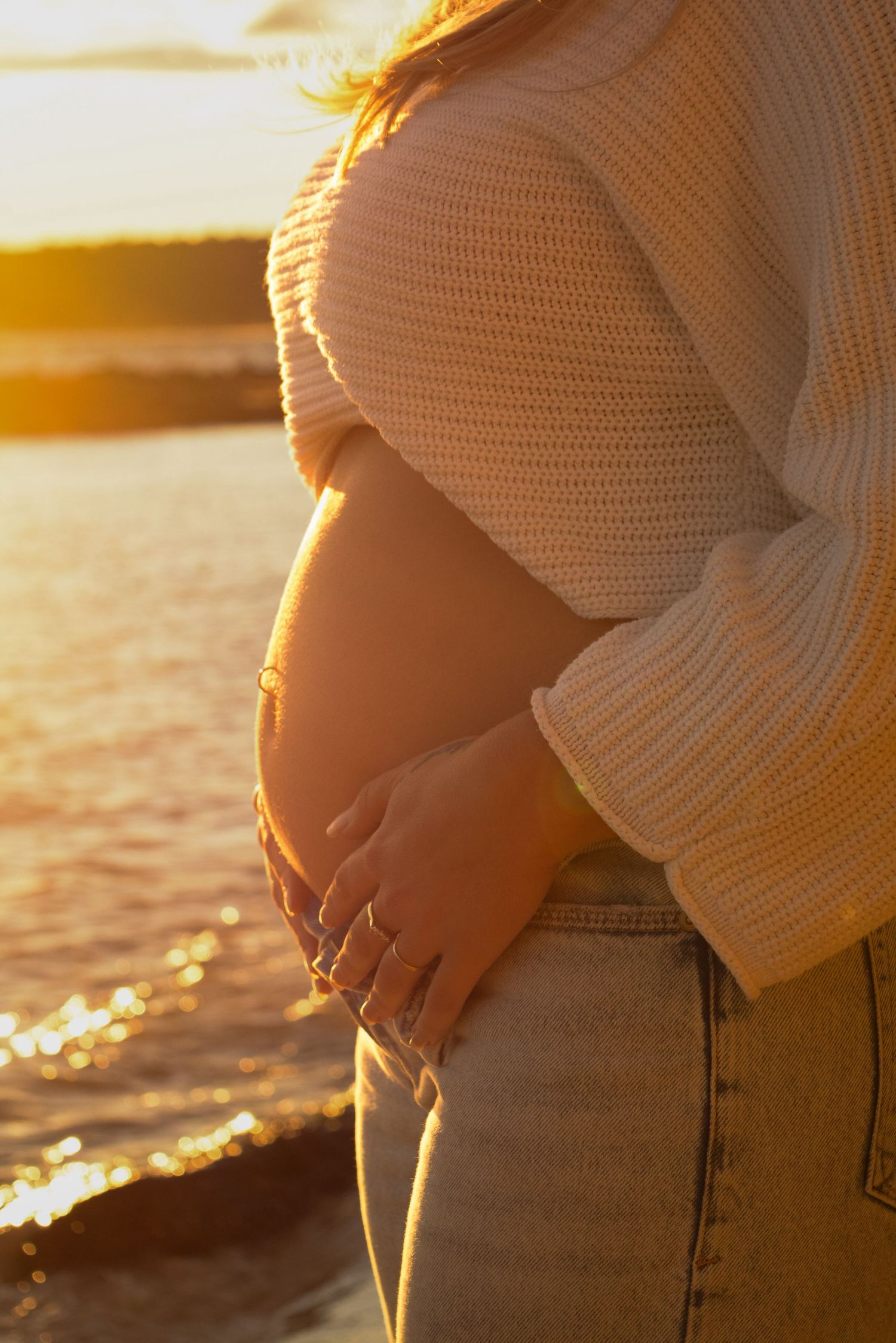 Side view of a pregnant belly in cream sweater illuminated by golden sunset light.