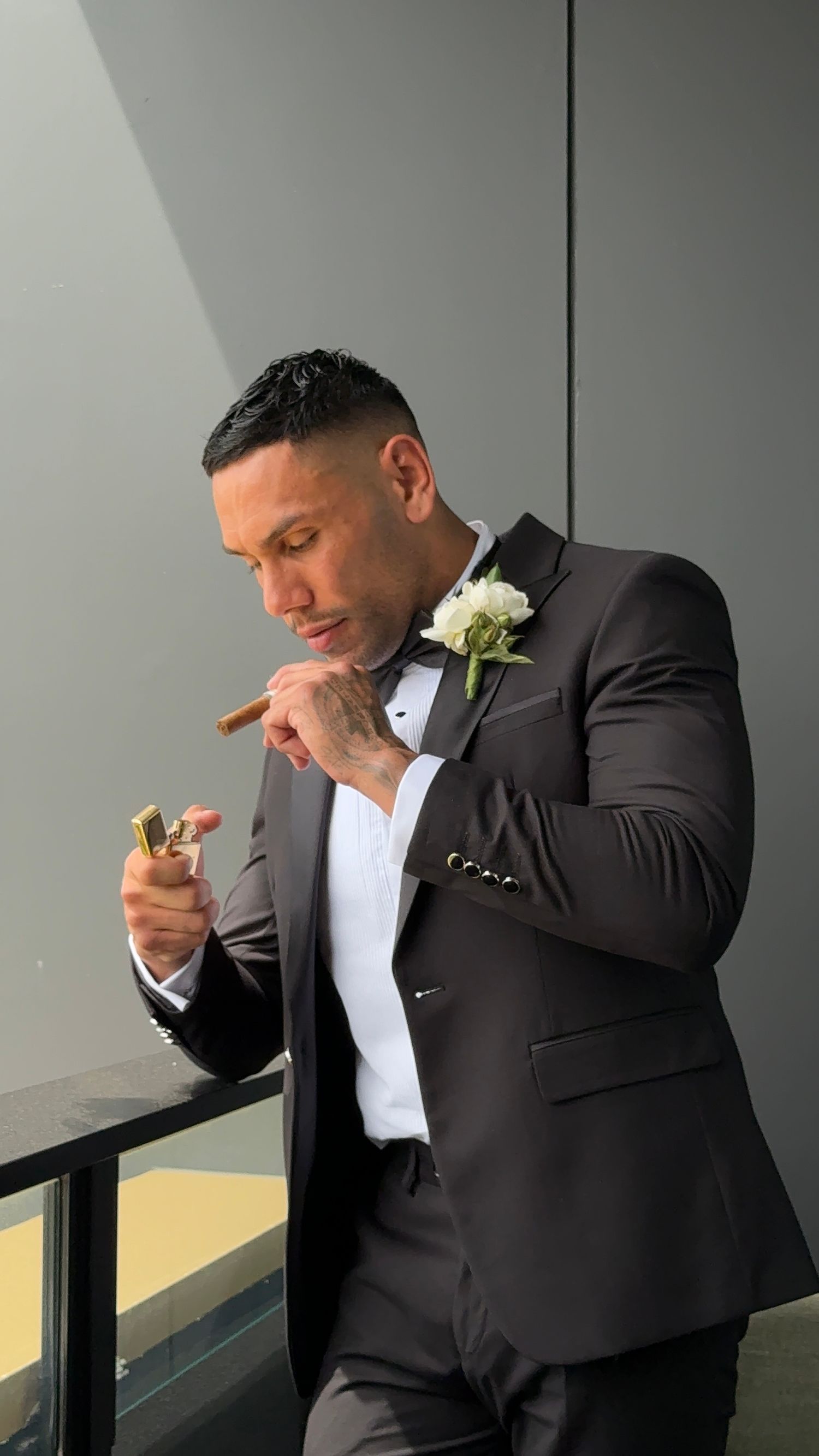 A person in a black suit and white shirt leans against a railing while lighting a cigar at a formal event.