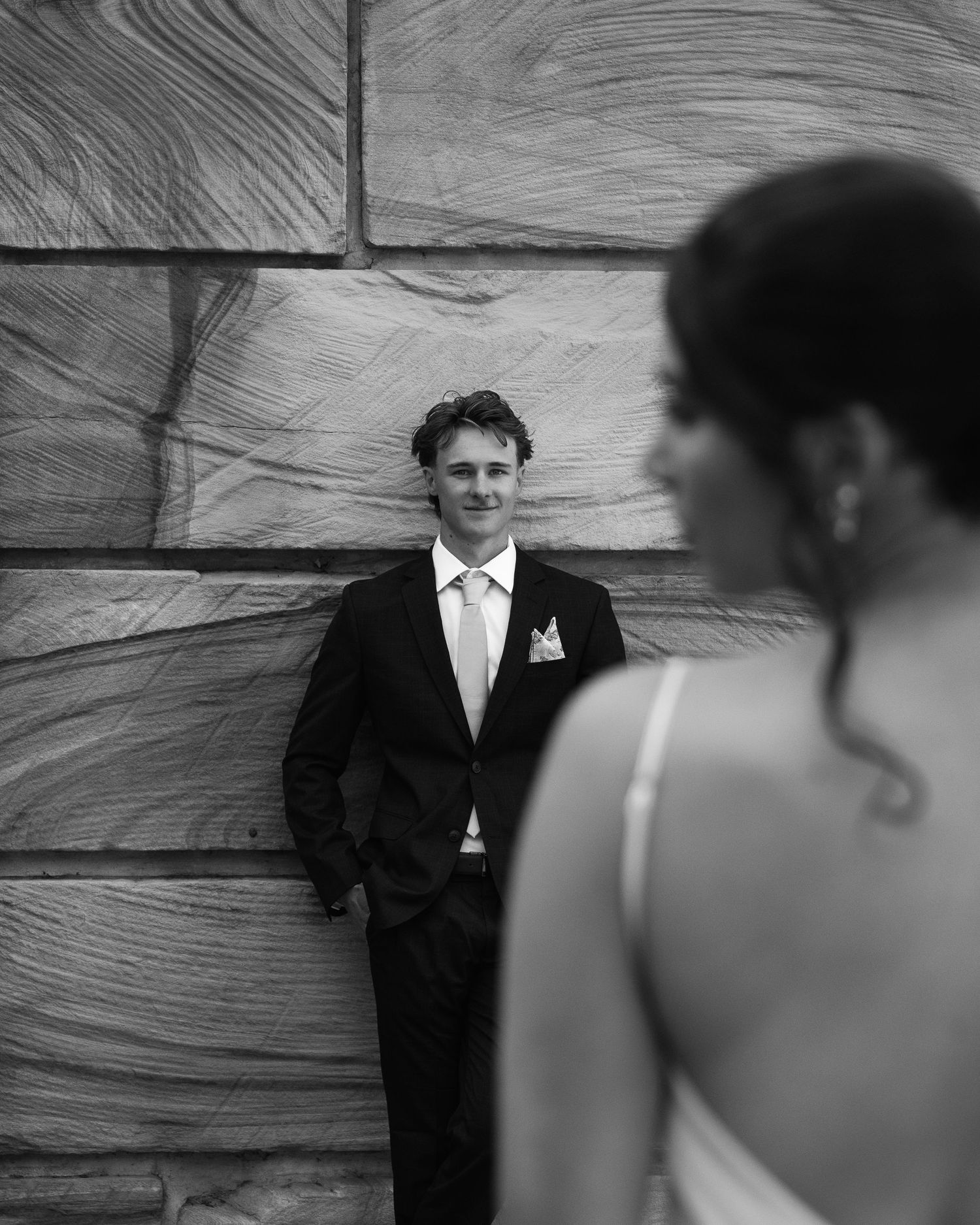 Black and white wedding photo of smiling groom in suit leaning against textured wall with bride in foreground.
