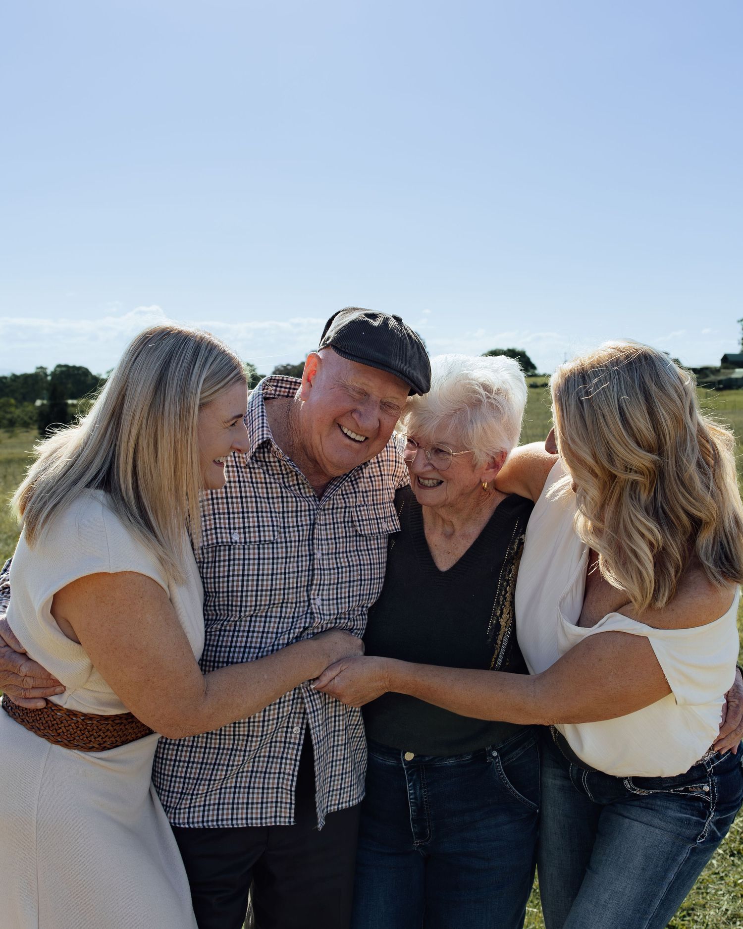 Four generations of family laughing and embracing outdoors on a sunny day in a green field.