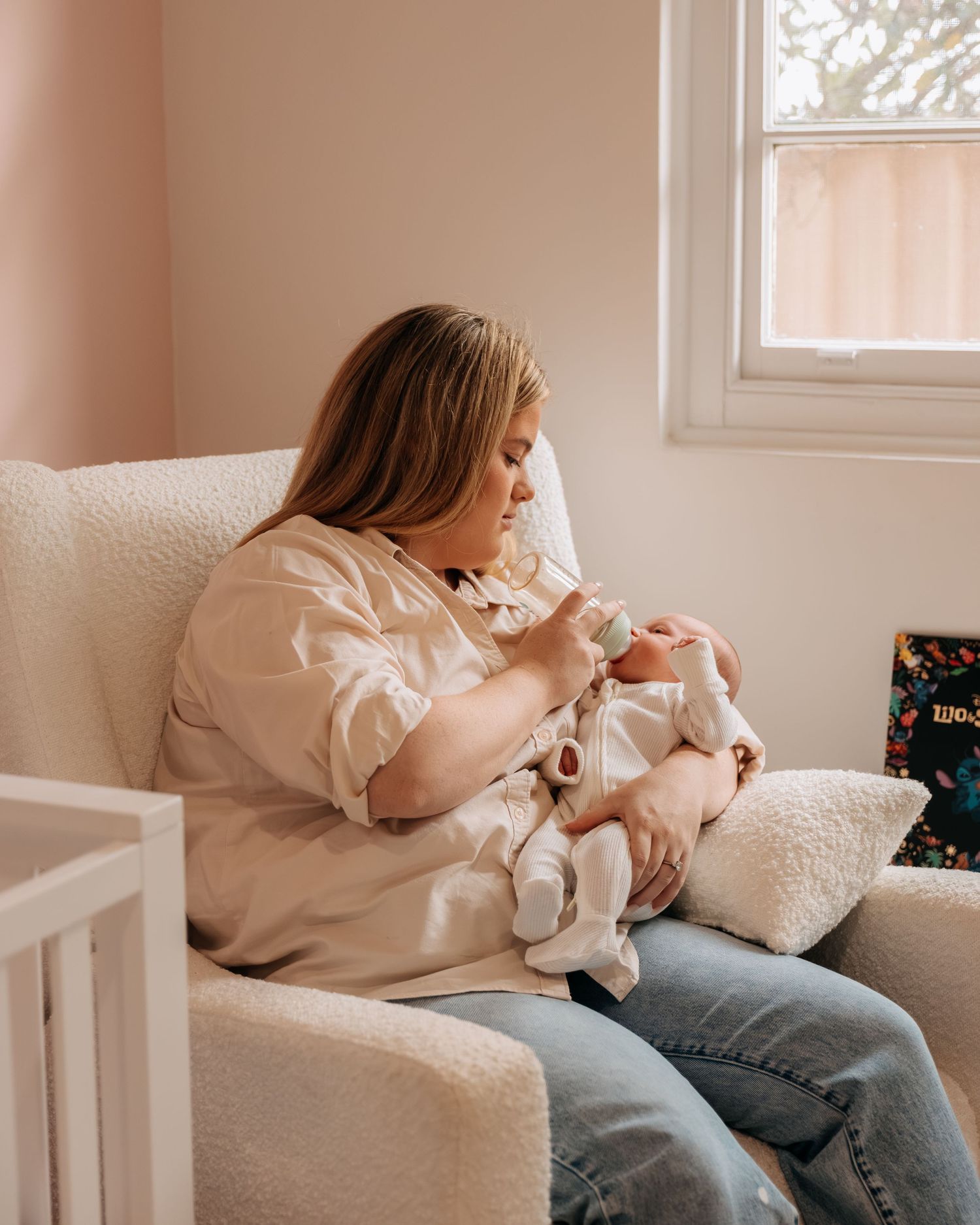 A mother sits in a cozy nursery chair bottle-feeding her newborn baby, wearing a white shirt and jeans.