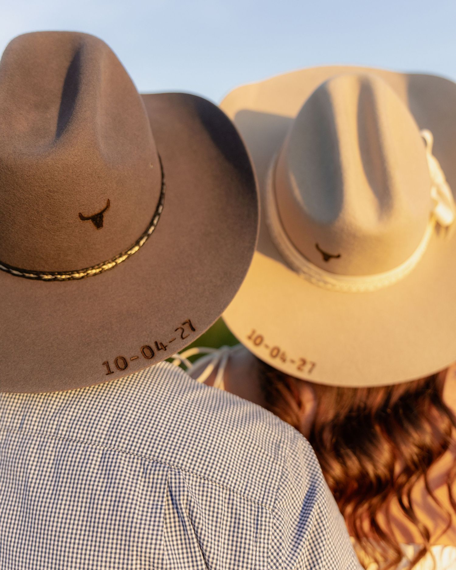 Couple wearing matching brown cowboy hats embroidered with a longhorn logo and date 10-04-27 from behind.