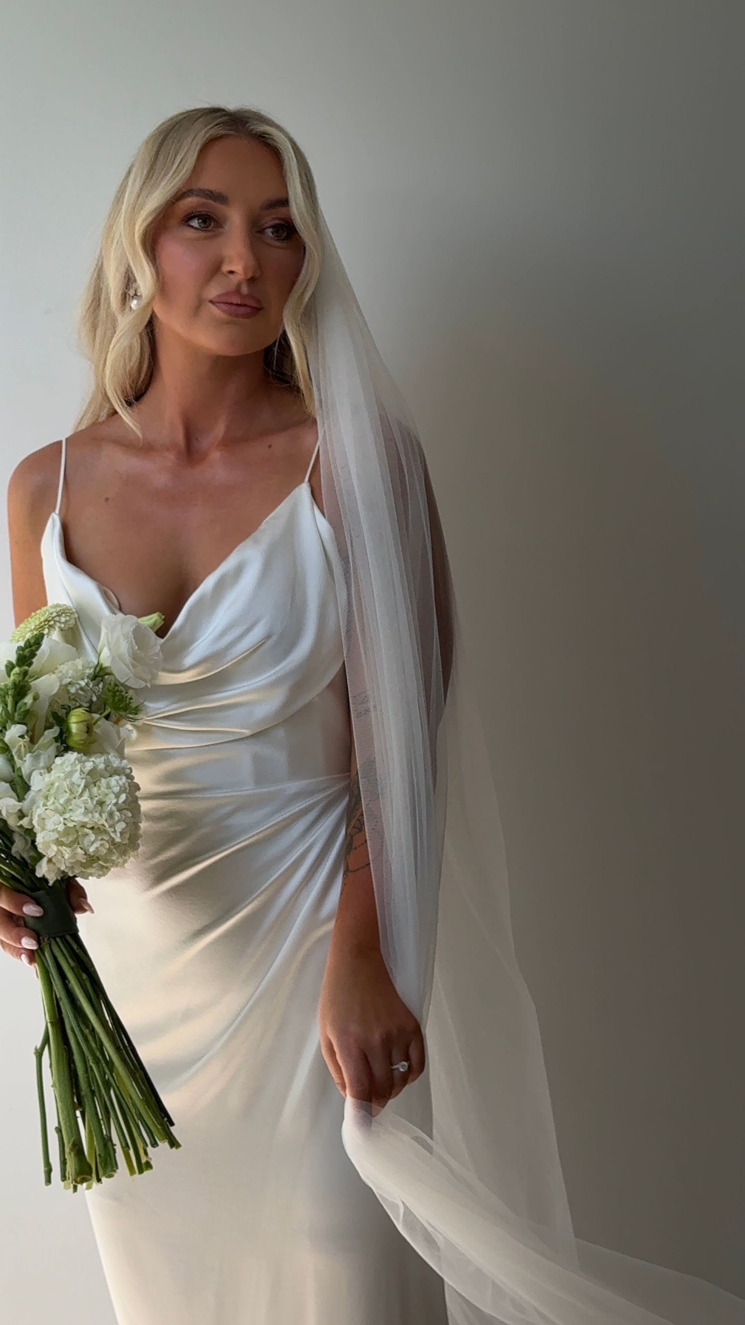 Bride in elegant white silk gown holds bouquet of white flowers while wearing flowing veil.