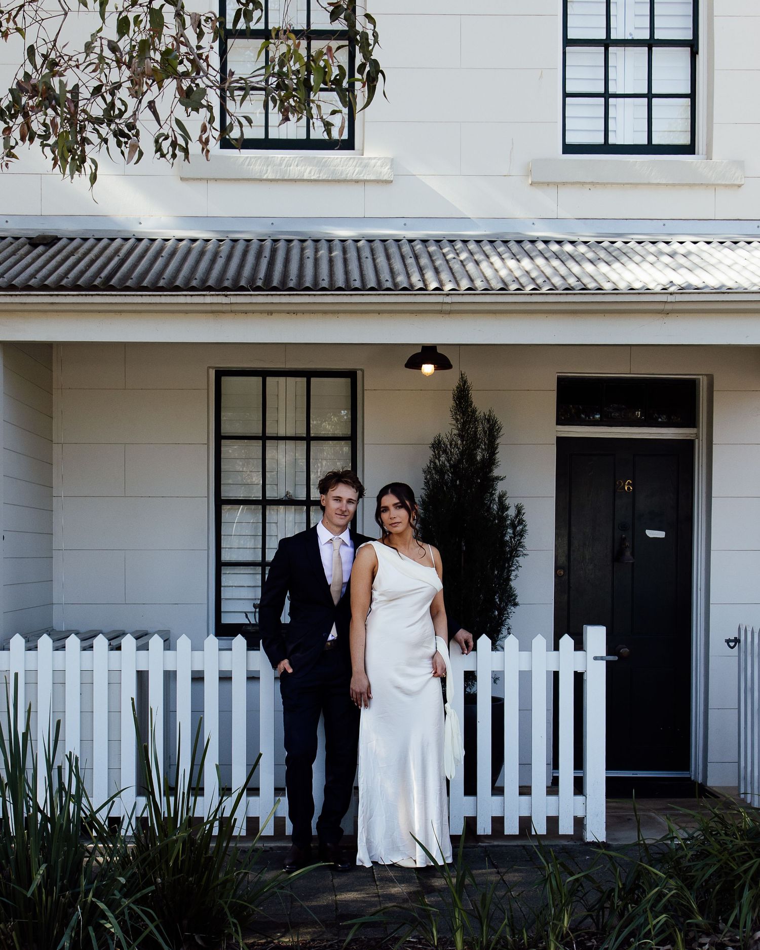 Couple in formal attire standing before a white picket fence outside a classic white home.