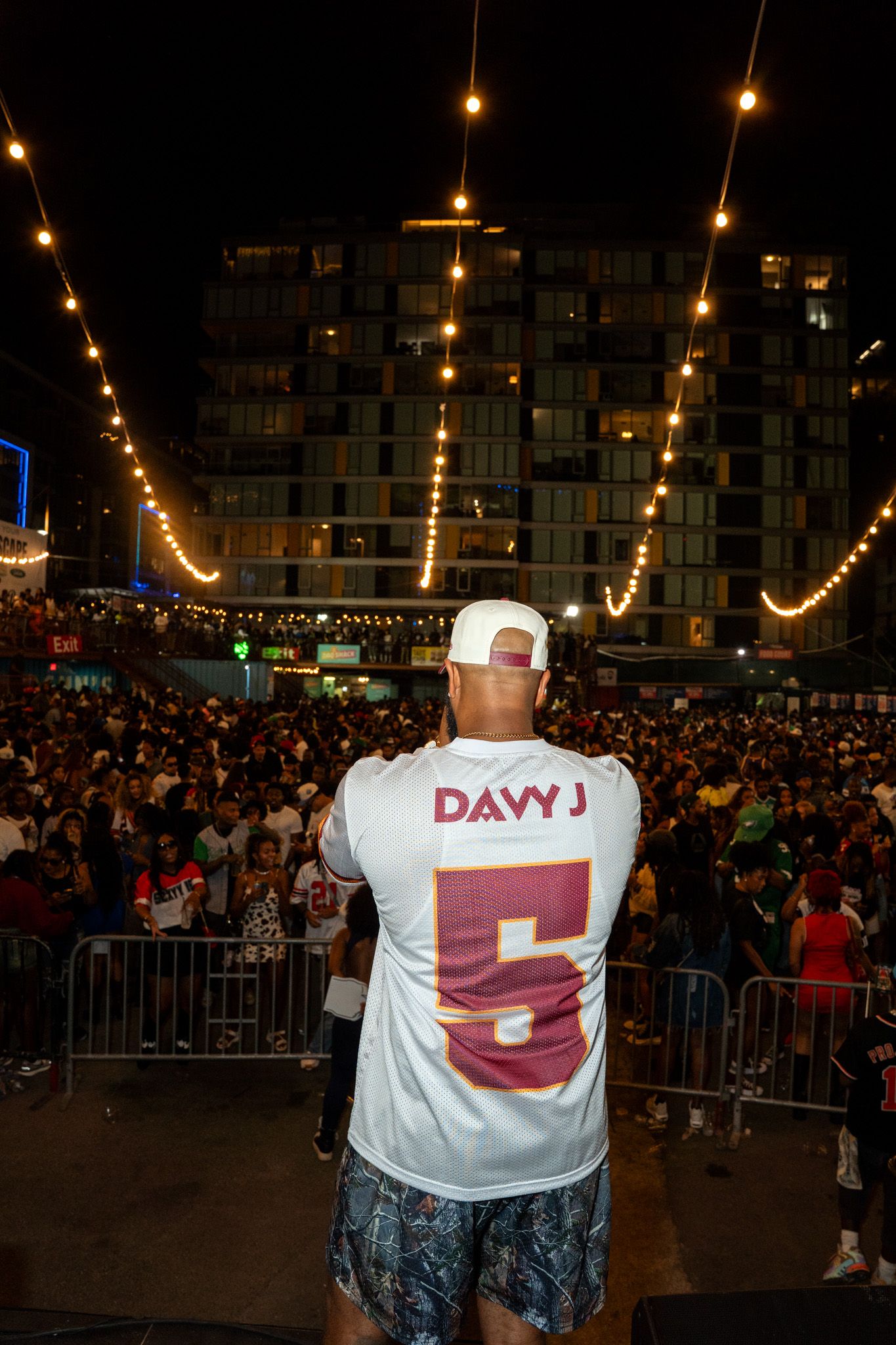 Nighttime crowd scene with string lights and person wearing white t-shirt with text.