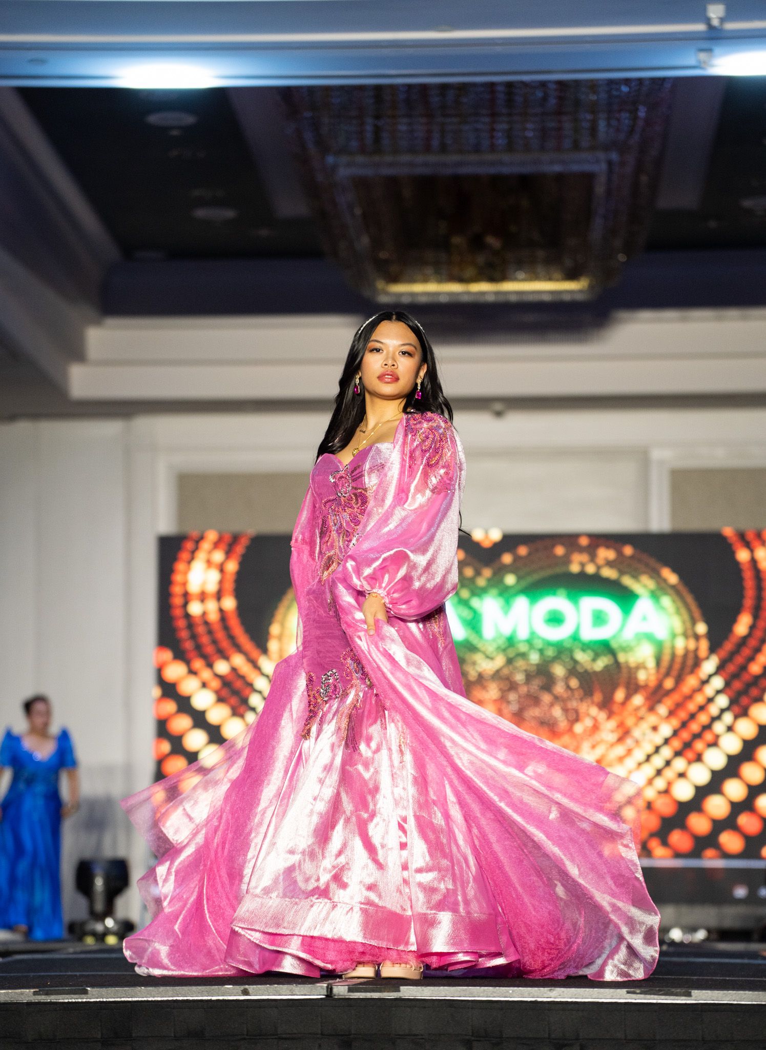 Model walks runway in flowing pink ball gown during fashion show with illuminated backdrop.