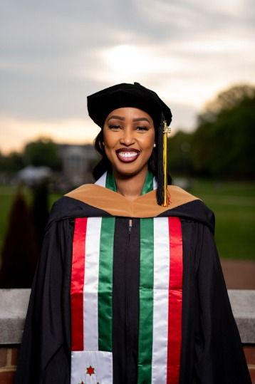 Graduate wearing traditional black academic regalia with colorful stoles poses outdoors at sunset.