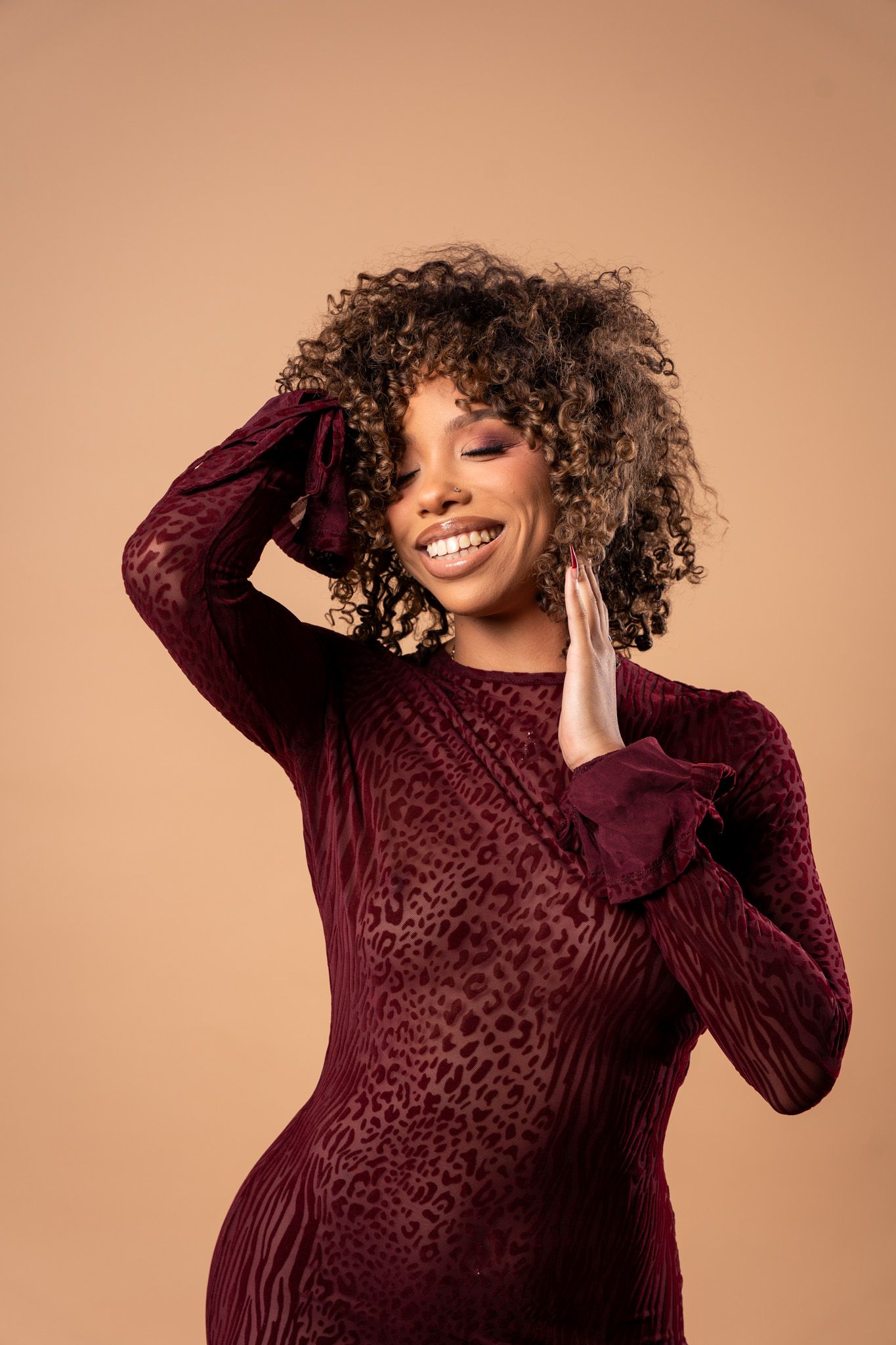 Smiling woman with curly hair wearing a burgundy animal print long-sleeve dress, posing against a beige background.