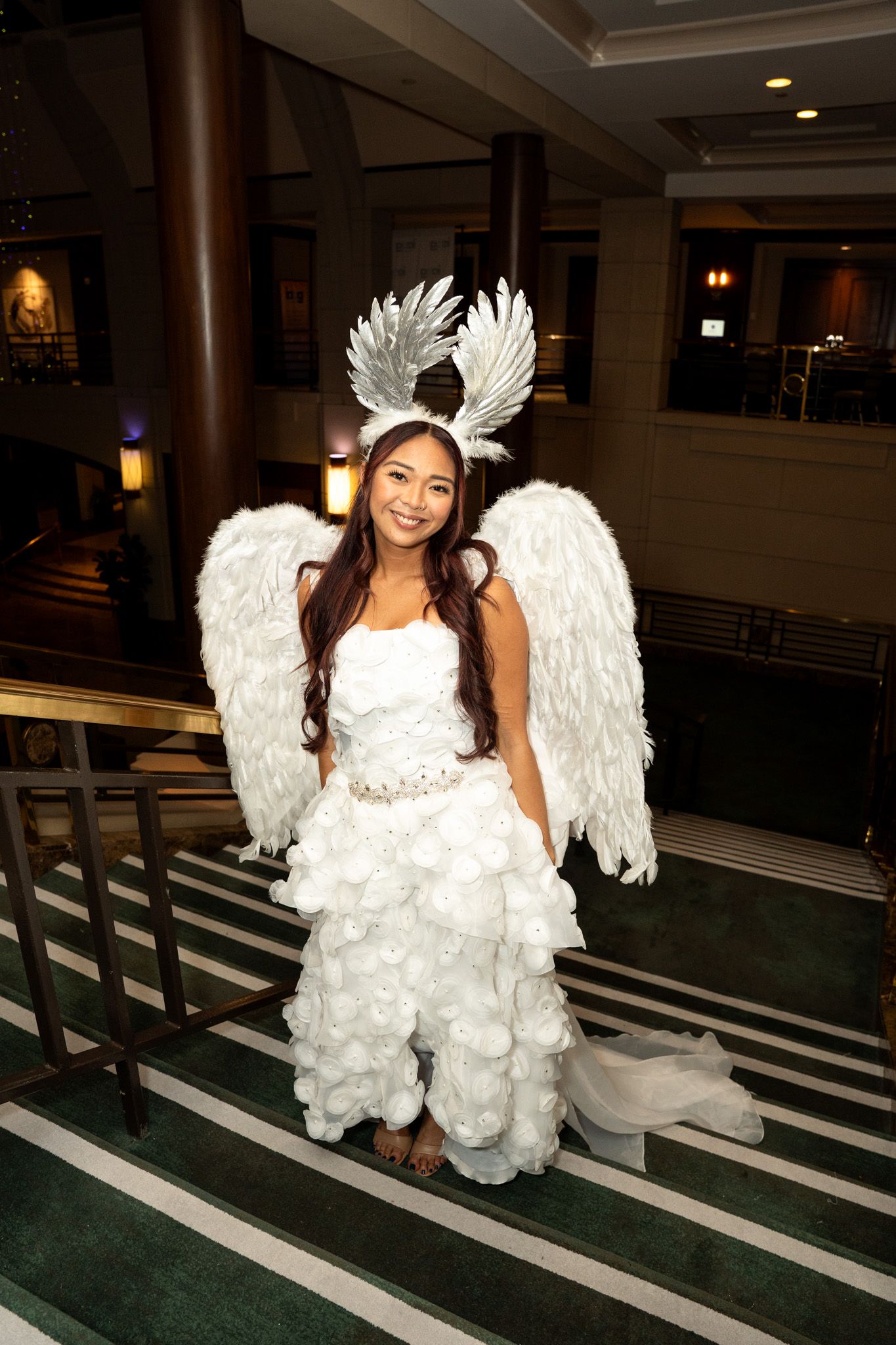 Model wearing a white feathered angel costume with wings poses in an elegant hallway.
