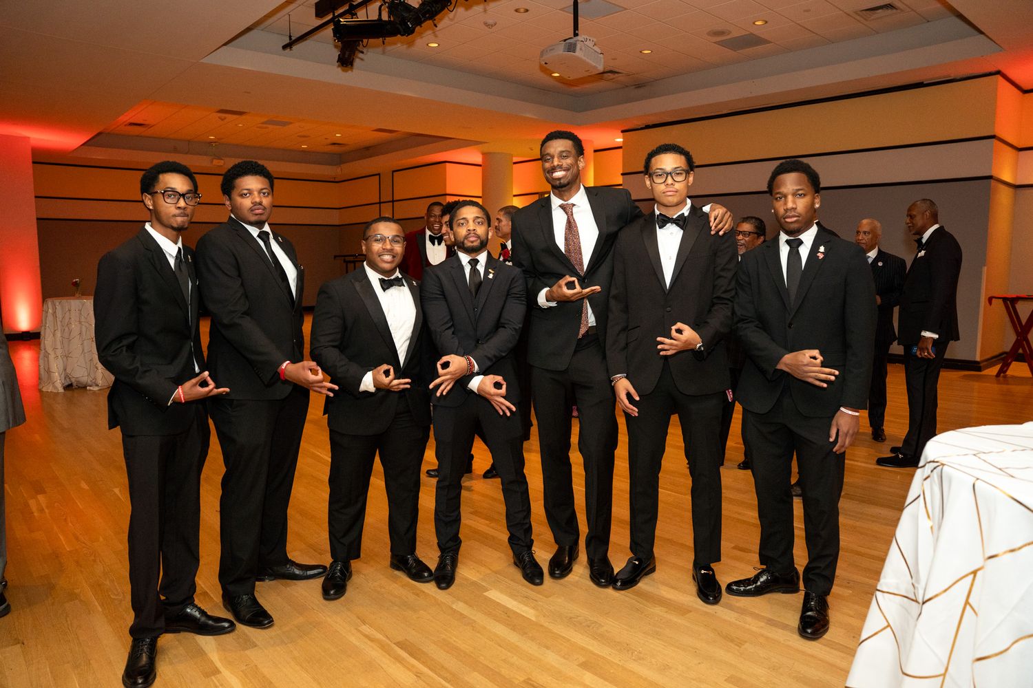 Group of men in formal black tuxedos pose together at elegant event with orange lighting.