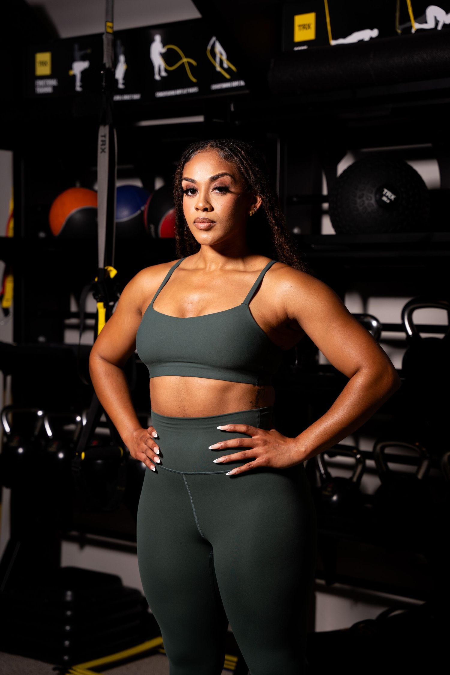 Fitness model posing in dark green athletic wear against gym equipment background.