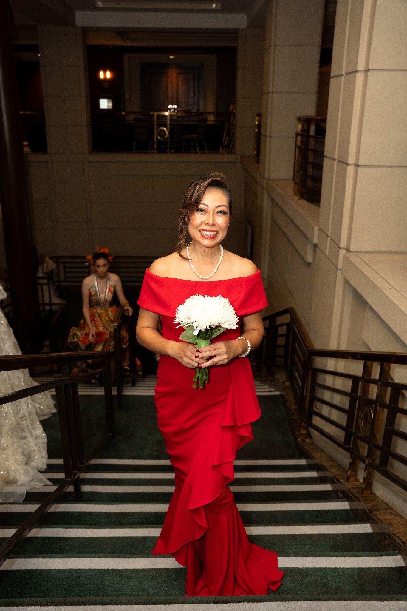 Model in flowing red off-shoulder gown poses with a bouquet in an elegant hallway.