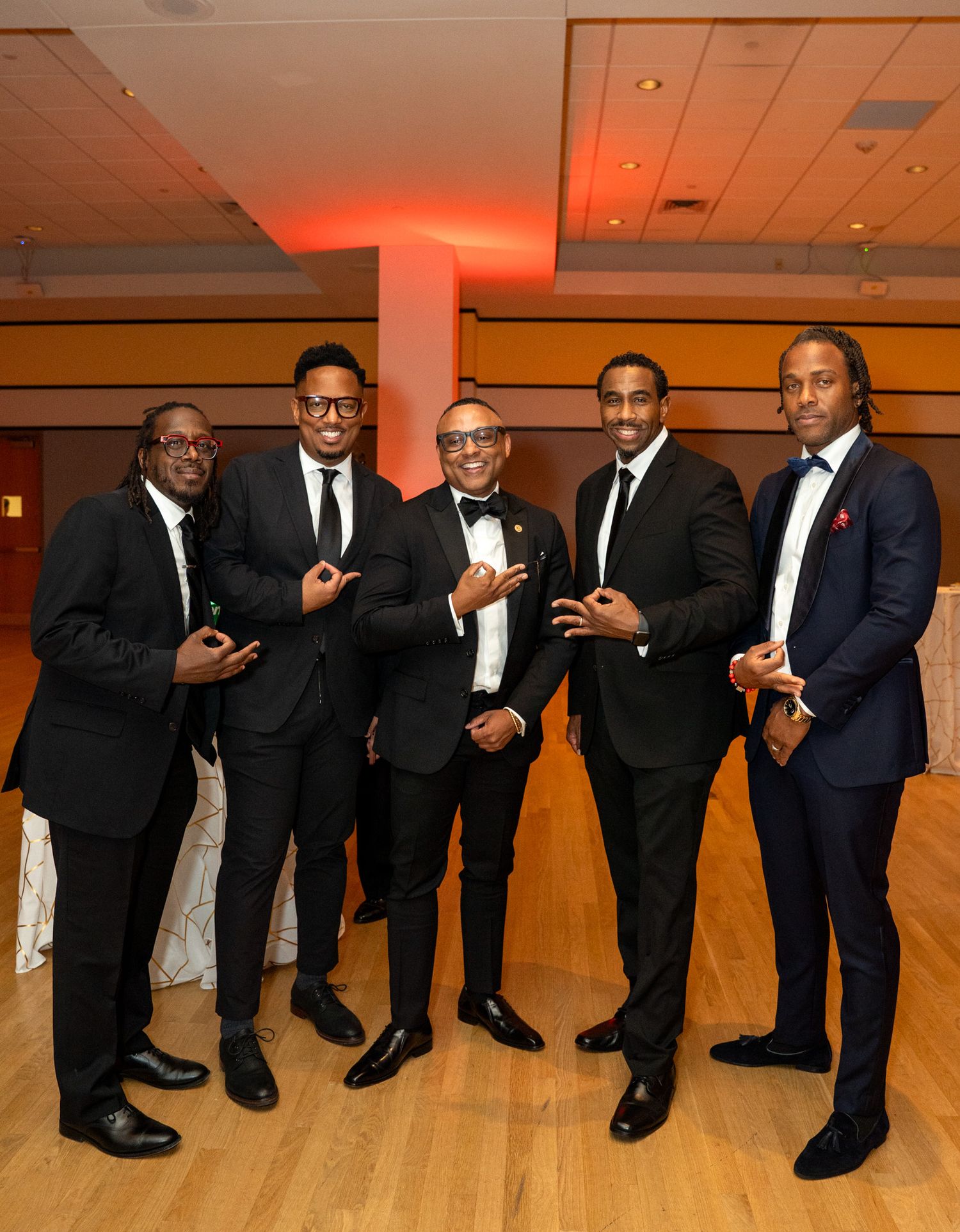 Five gentlemen in black tuxedos pose together at formal event with orange accent lighting.