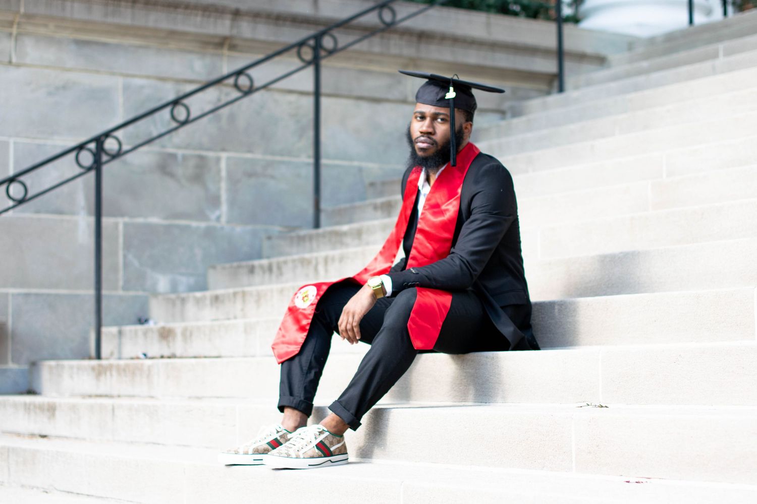 Graduate in black suit and red stole sits on concrete steps for graduation photos.