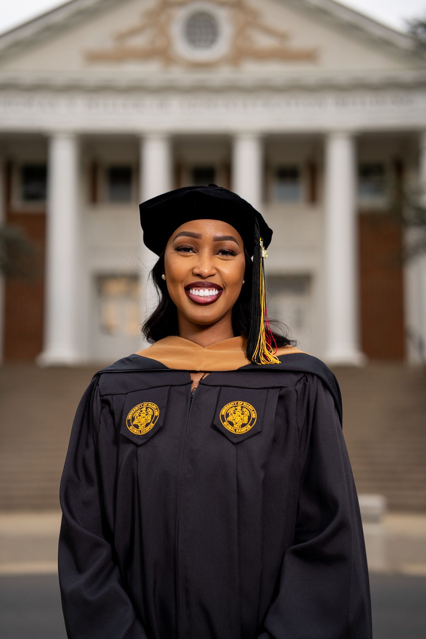 Graduate in traditional black academic regalia stands before columned building.