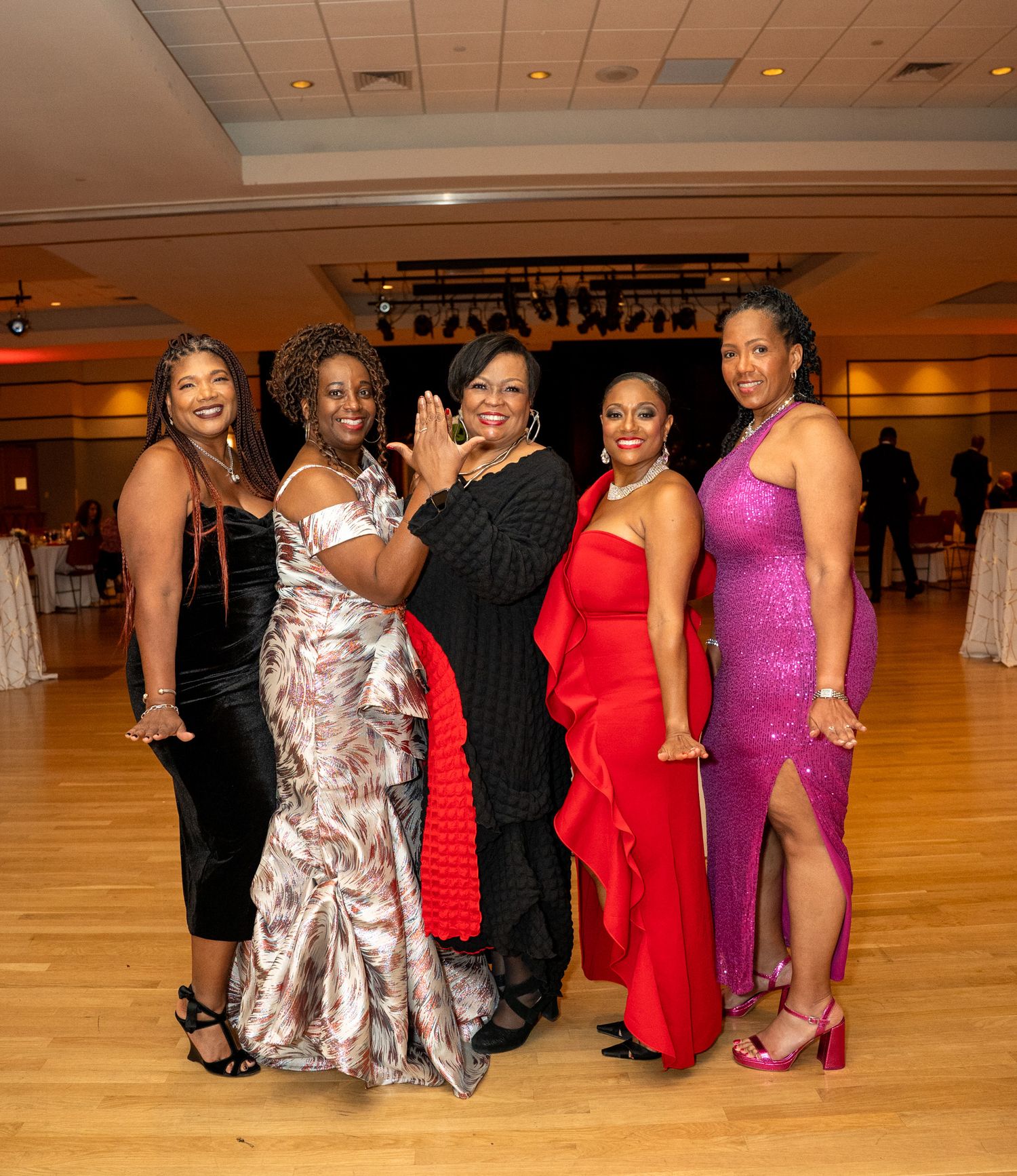 Group of women in elegant evening gowns pose together at formal event with wooden dance floor.