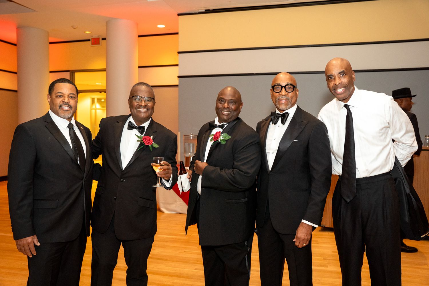 Five men in formal black tie attire smile during an evening event with warm ambient lighting.