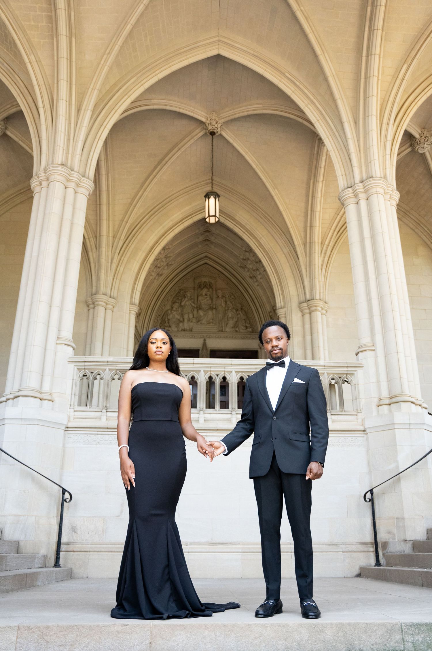 Formal couple stands holding hands under Gothic arches in elegant architectural setting.