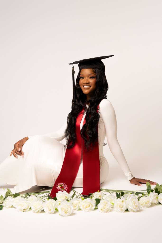 Graduate poses with white roses in elegant red and white graduation attire against light background.
