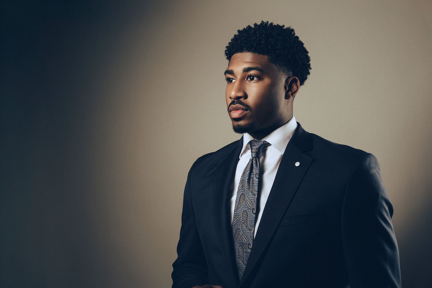 Professional portrait of someone in a dark suit and patterned tie against a moody gradient background.