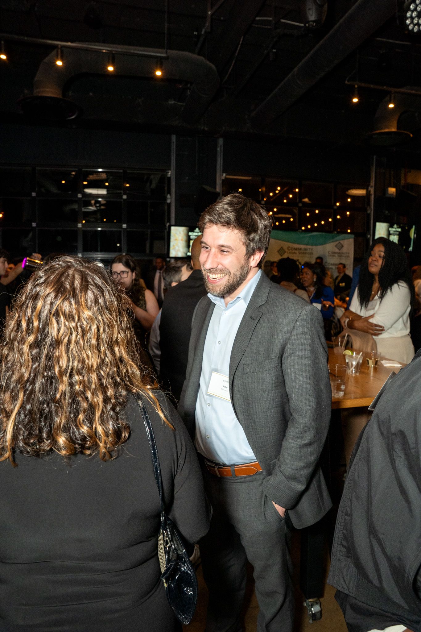 People engaged in conversation at an indoor event with mood lighting and exposed ceiling.