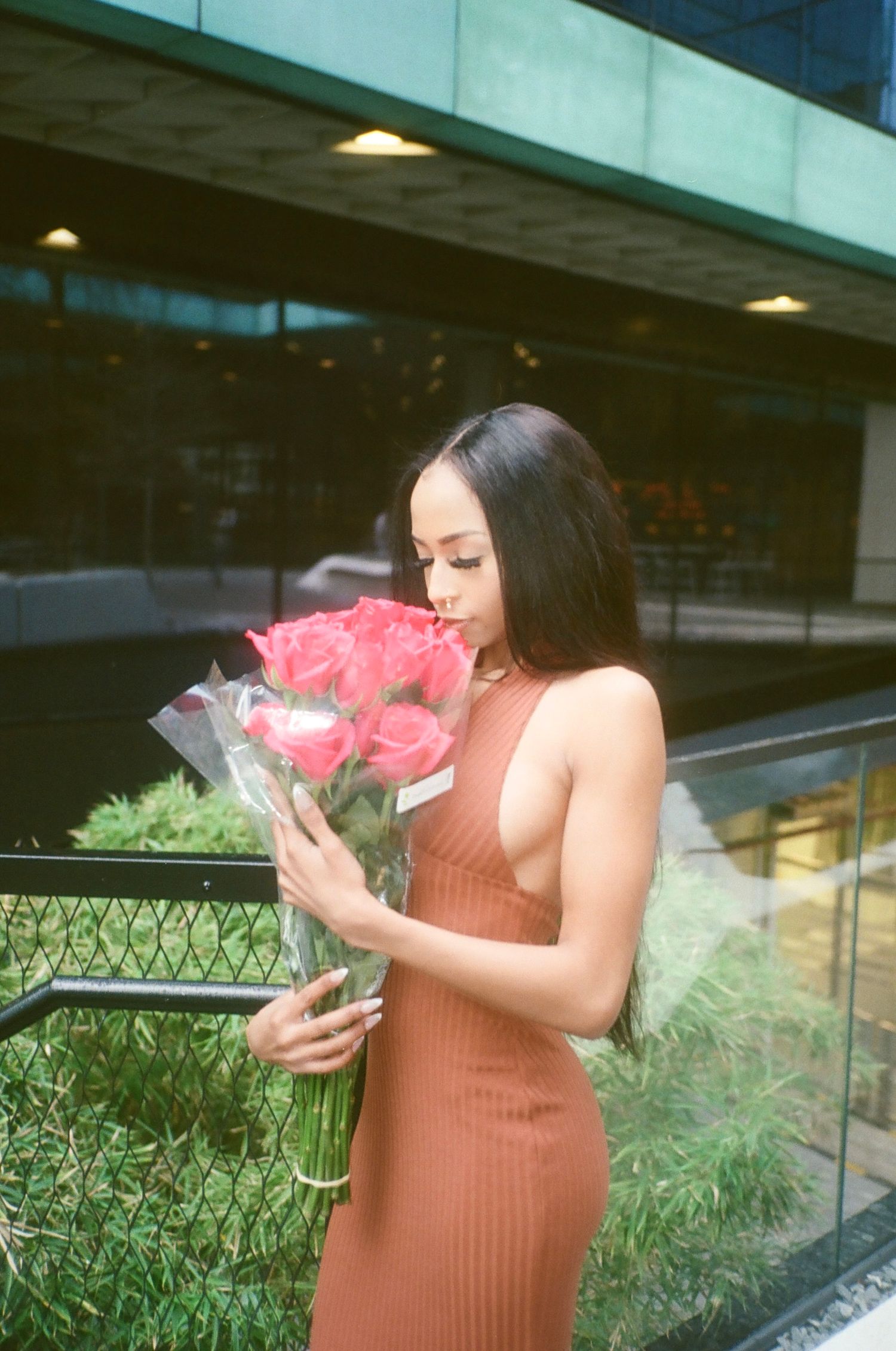 Person in coral dress holding pink roses bouquet outside modern building.
