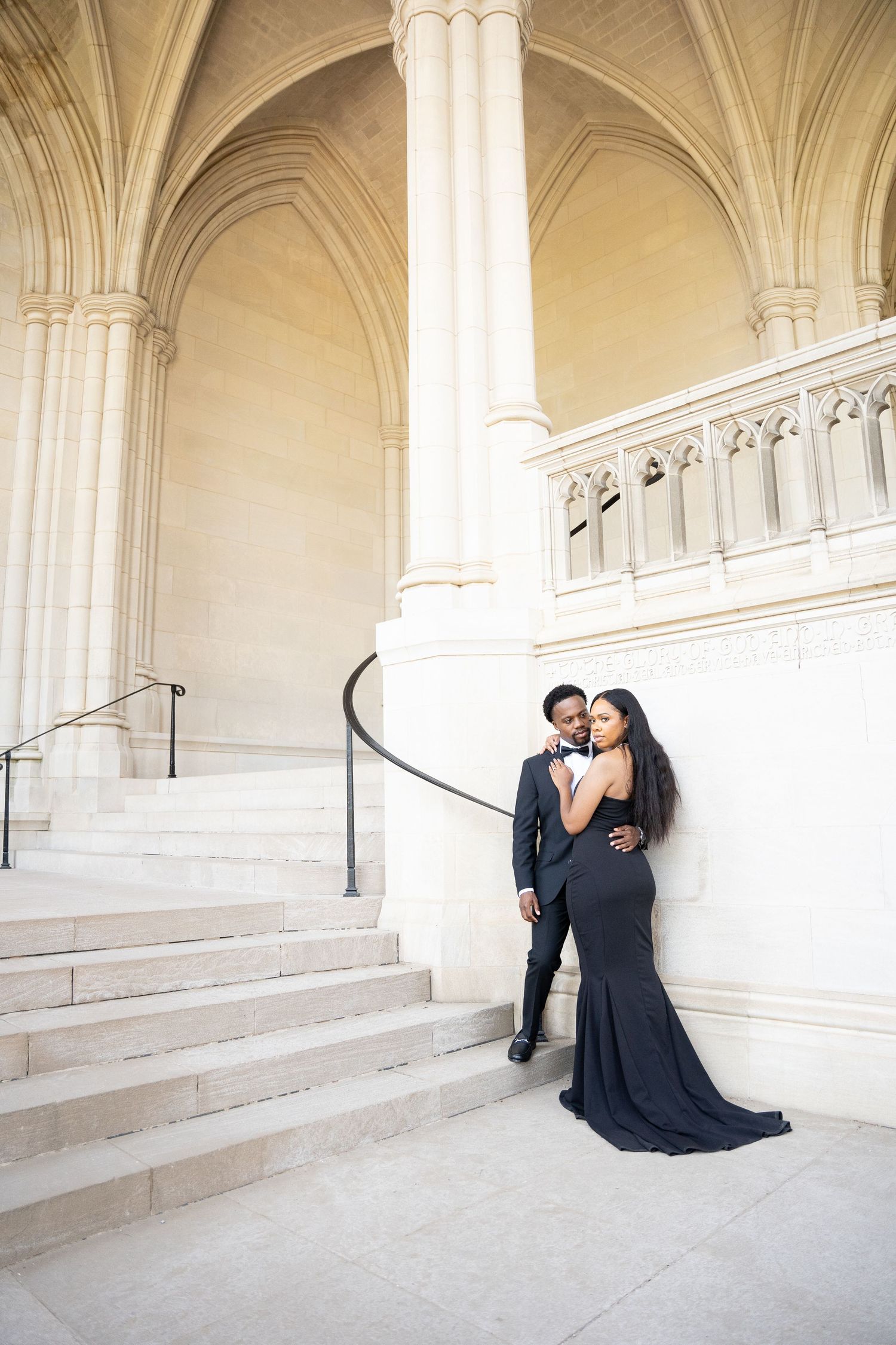 Couple in formal wear embracing on grand white marble staircase with vaulted ceiling.