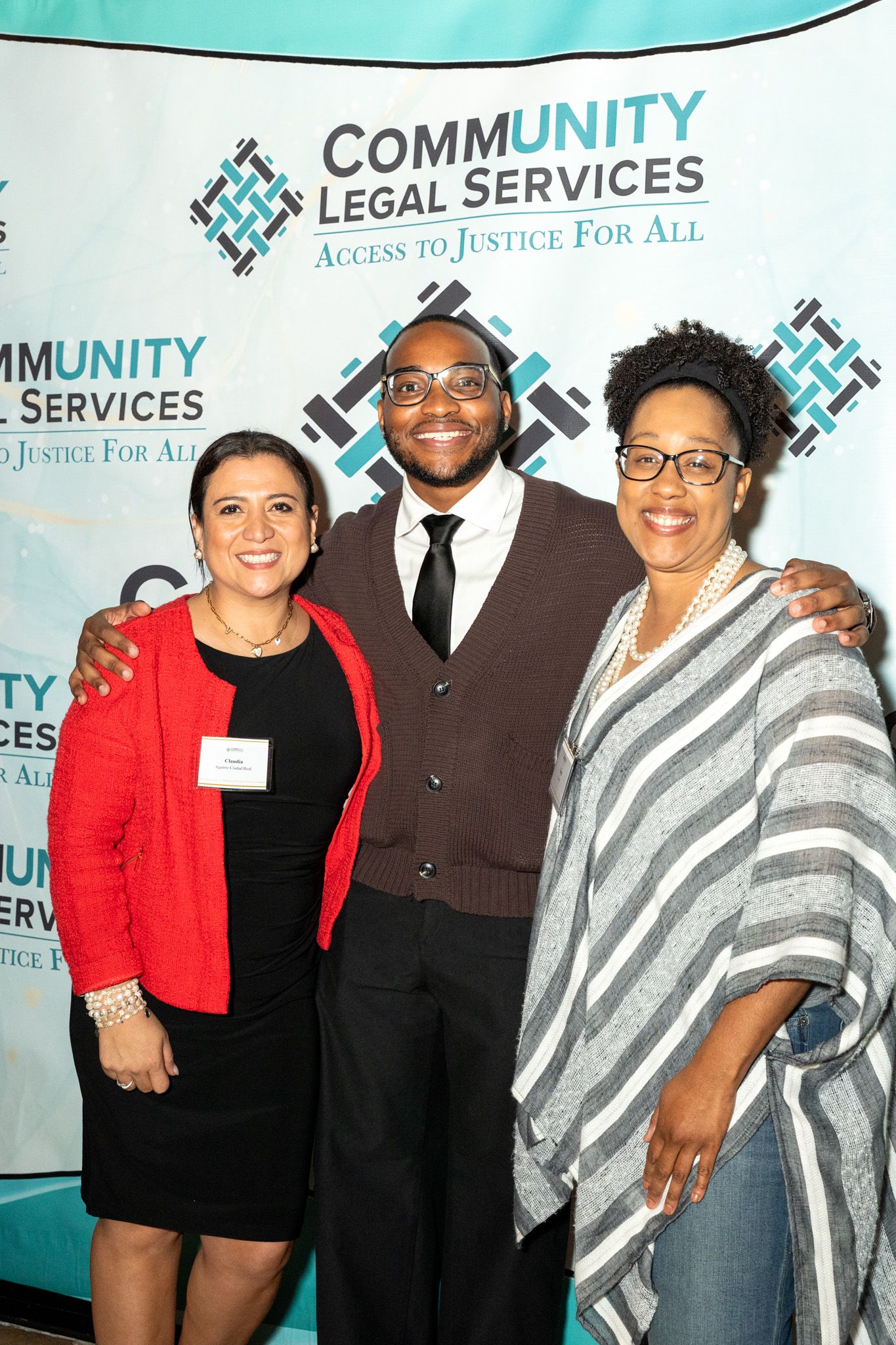 A group of people pose together at a Community Legal Services event with a teal backdrop and organization logos.