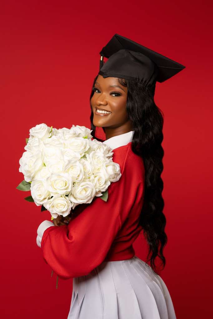 Graduate in red and white attire holds bouquet of white roses against red background during graduation photoshoot.