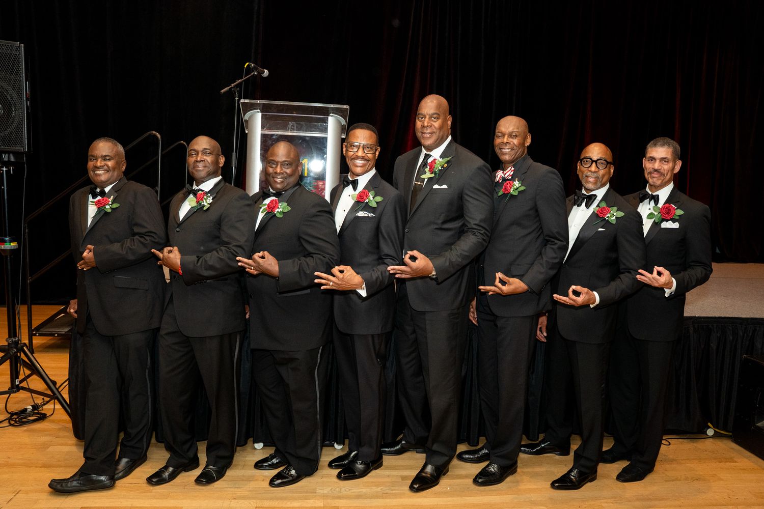 Row of men in formal black suits pose together on stage during special event.