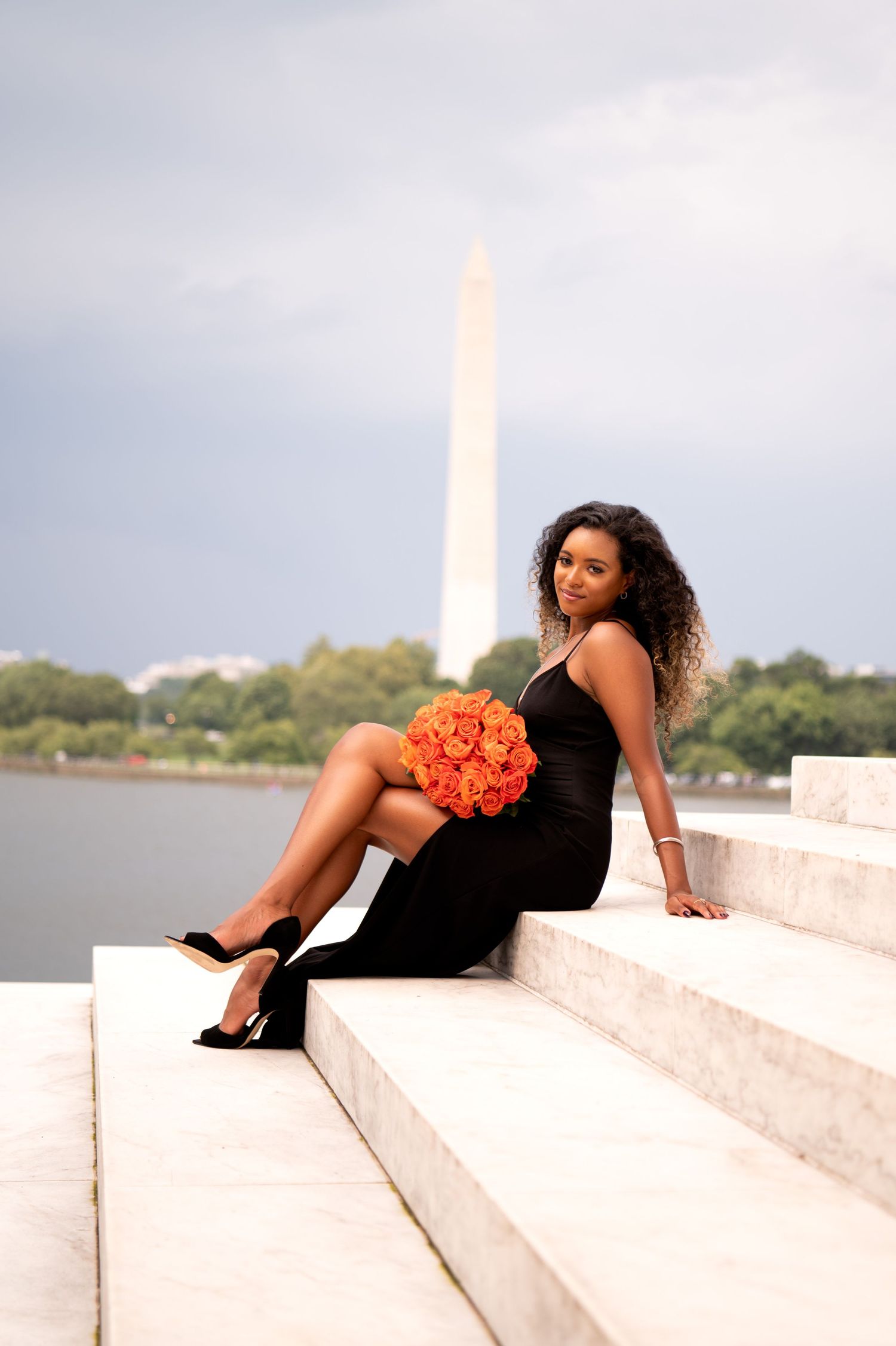 Portrait session on steps with black dress and orange flowers near water.