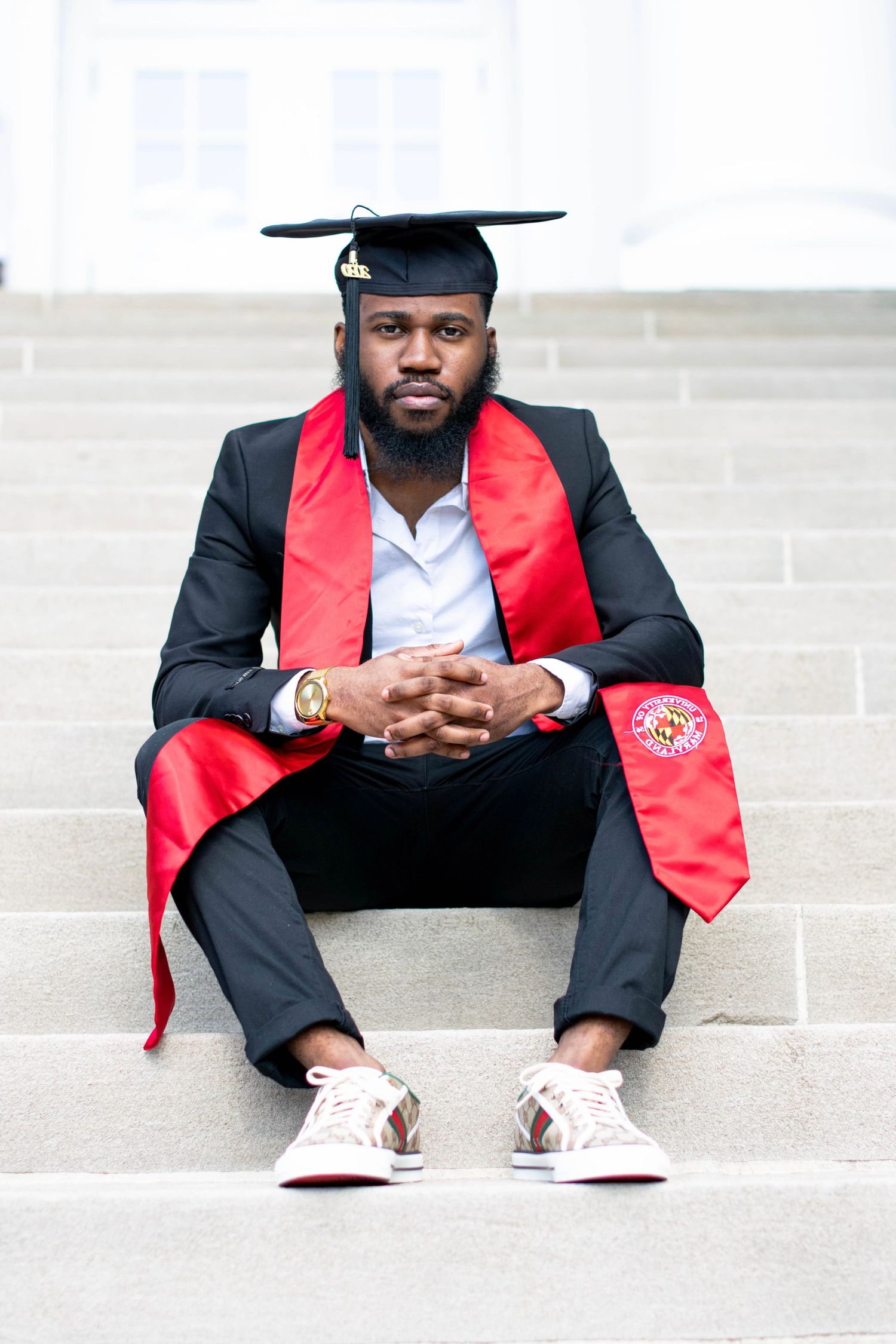 Graduate in formal attire and red stole poses on steps during graduation photoshoot.