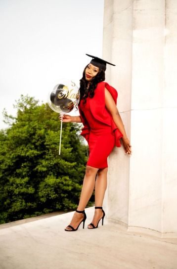 Graduate in red dress and cap poses with silver balloon against white pillar outdoors.