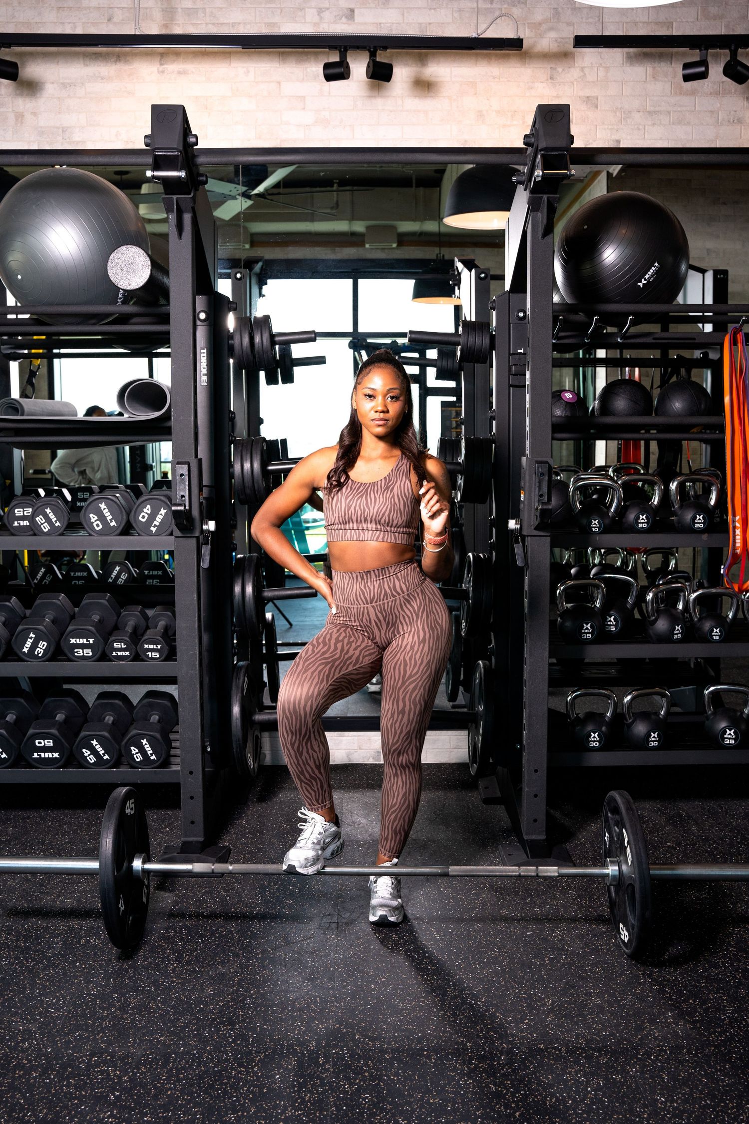 Athlete in brown workout gear posing between exercise equipment in bright gym setting.