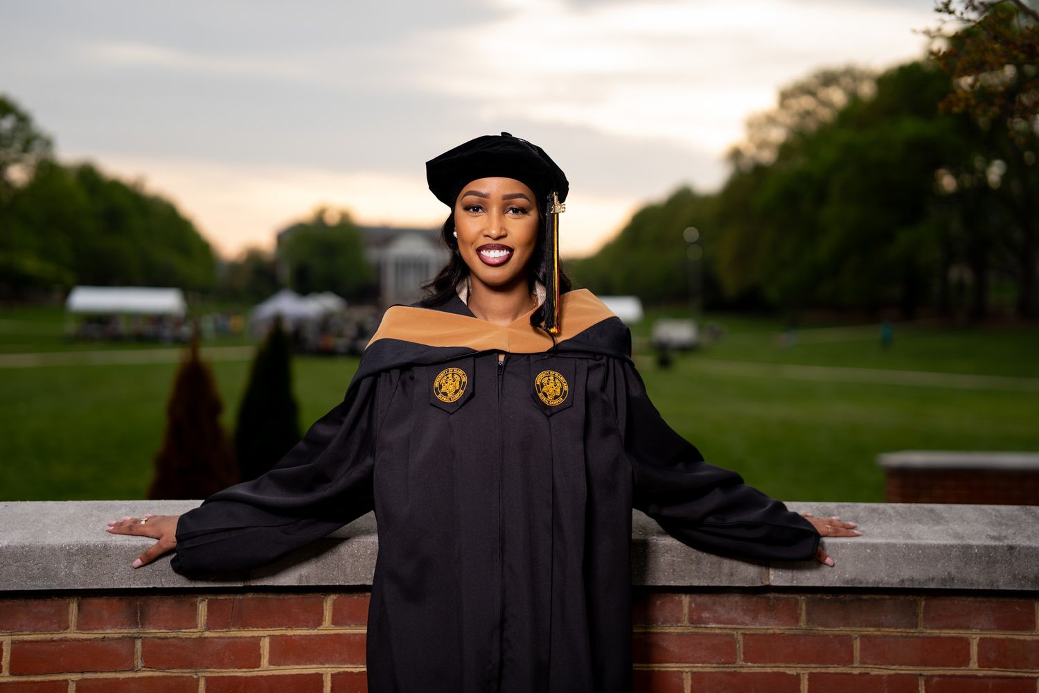 Graduate in black academic gown with decorative emblems poses on brick wall.