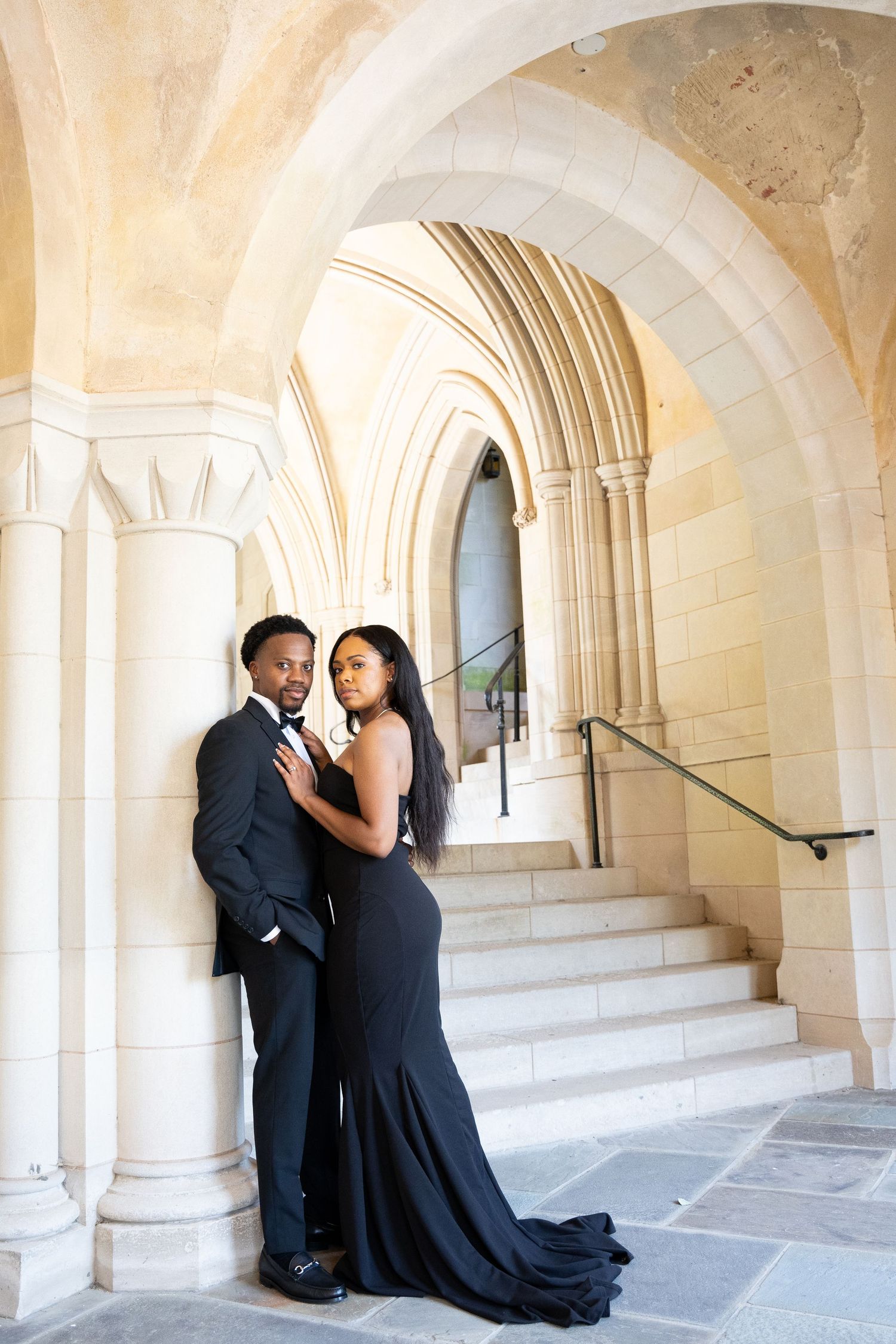 Elegant couple poses in formal attire against Gothic archway and stone staircase.