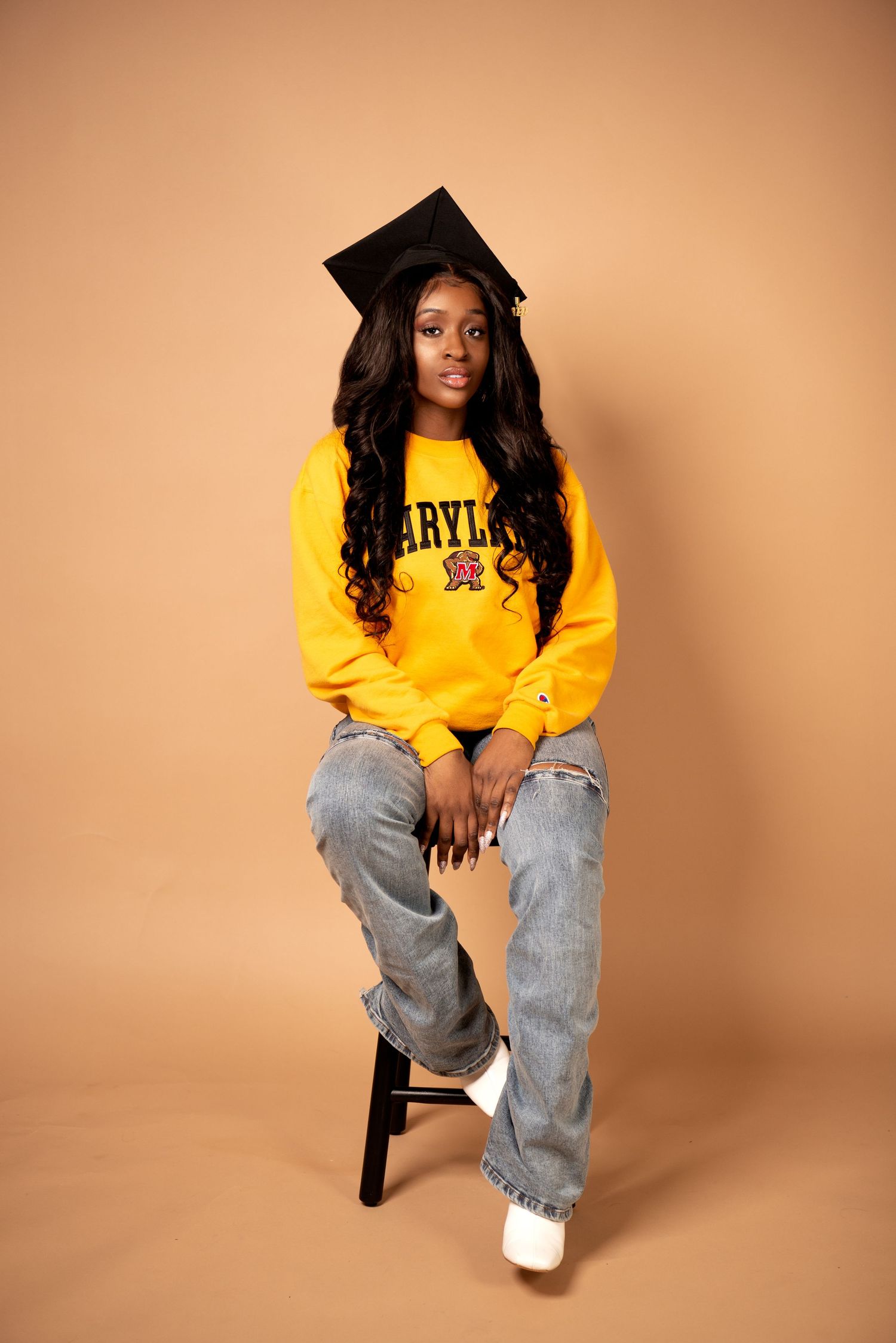 Graduate in yellow university sweatshirt and cap poses against warm backdrop.