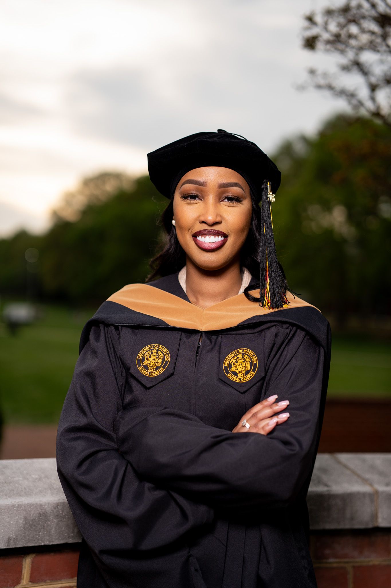 Graduate in doctoral regalia with gold emblems poses outdoors at sunset.