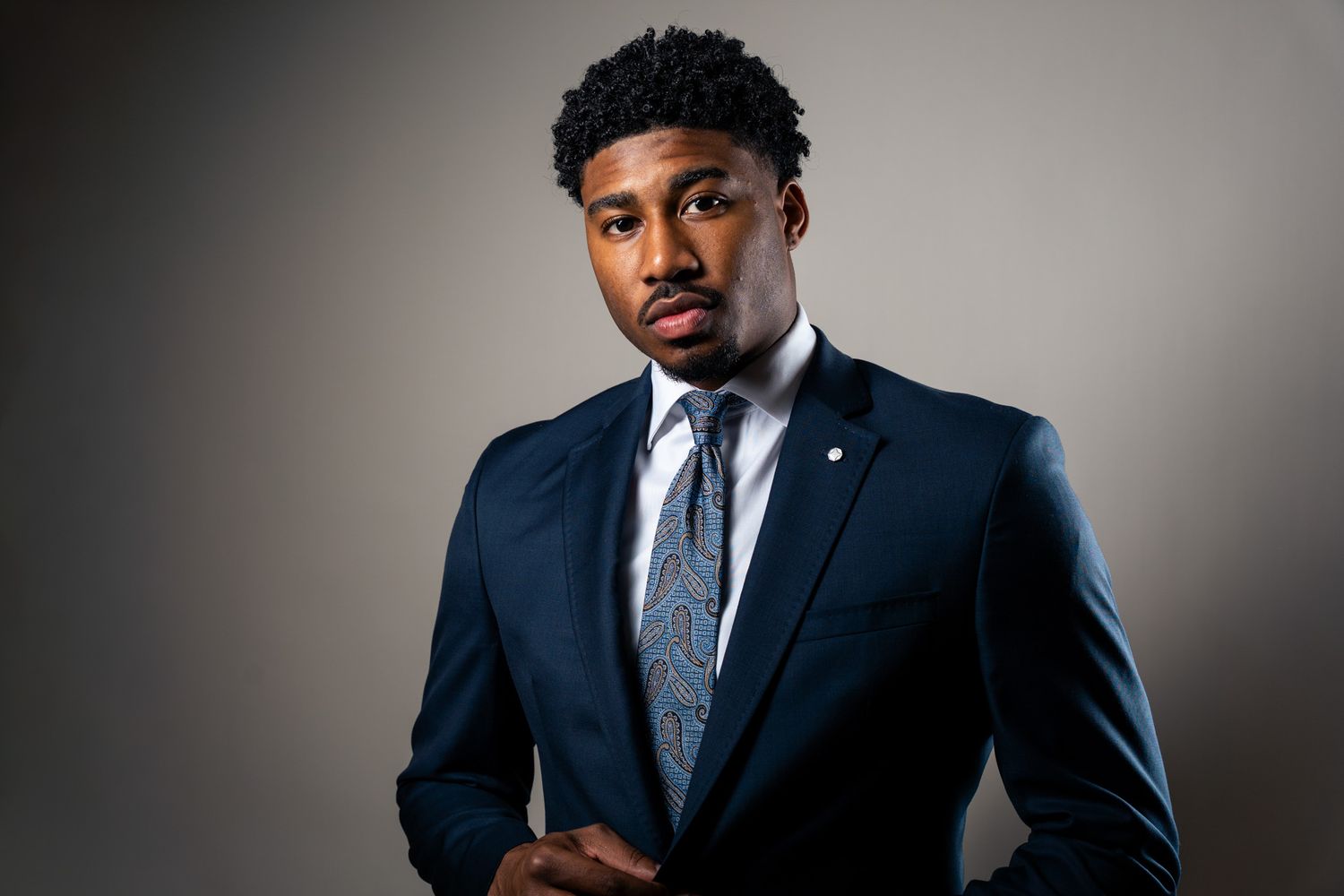 Professional portrait of a person wearing a navy blue suit and silver tie against a gray studio backdrop.