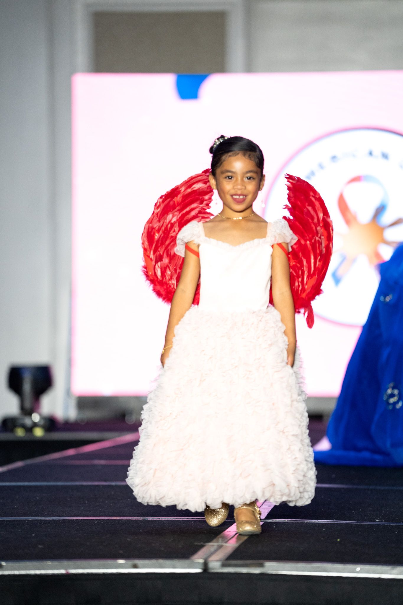 Young performer in white dress with red wings dances on dark runway stage.