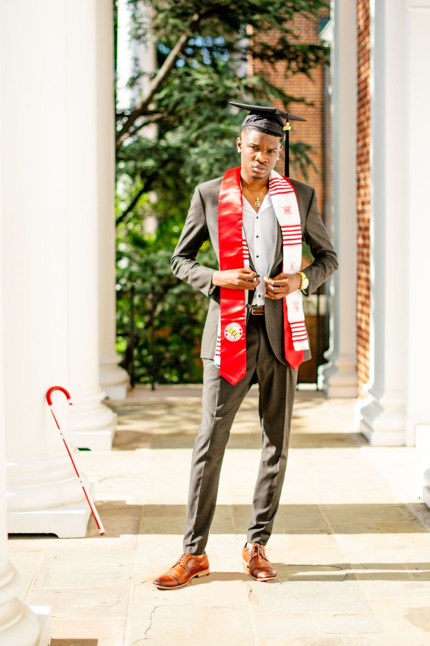 Graduate in grey suit and red stole poses near white columns on graduation day.