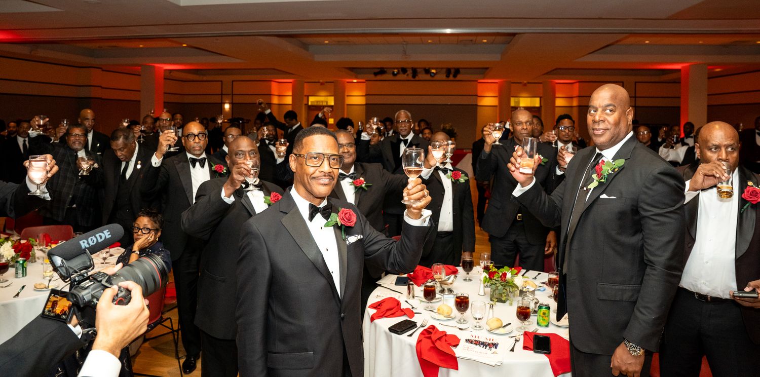 Formal event in a banquet hall with guests in black tie attire at dining tables.