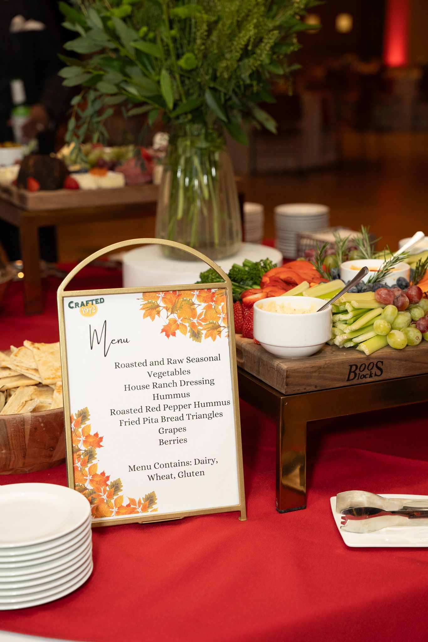 Buffet table with autumn-themed menu card, fresh vegetables, and appetizer spread on red tablecloth.